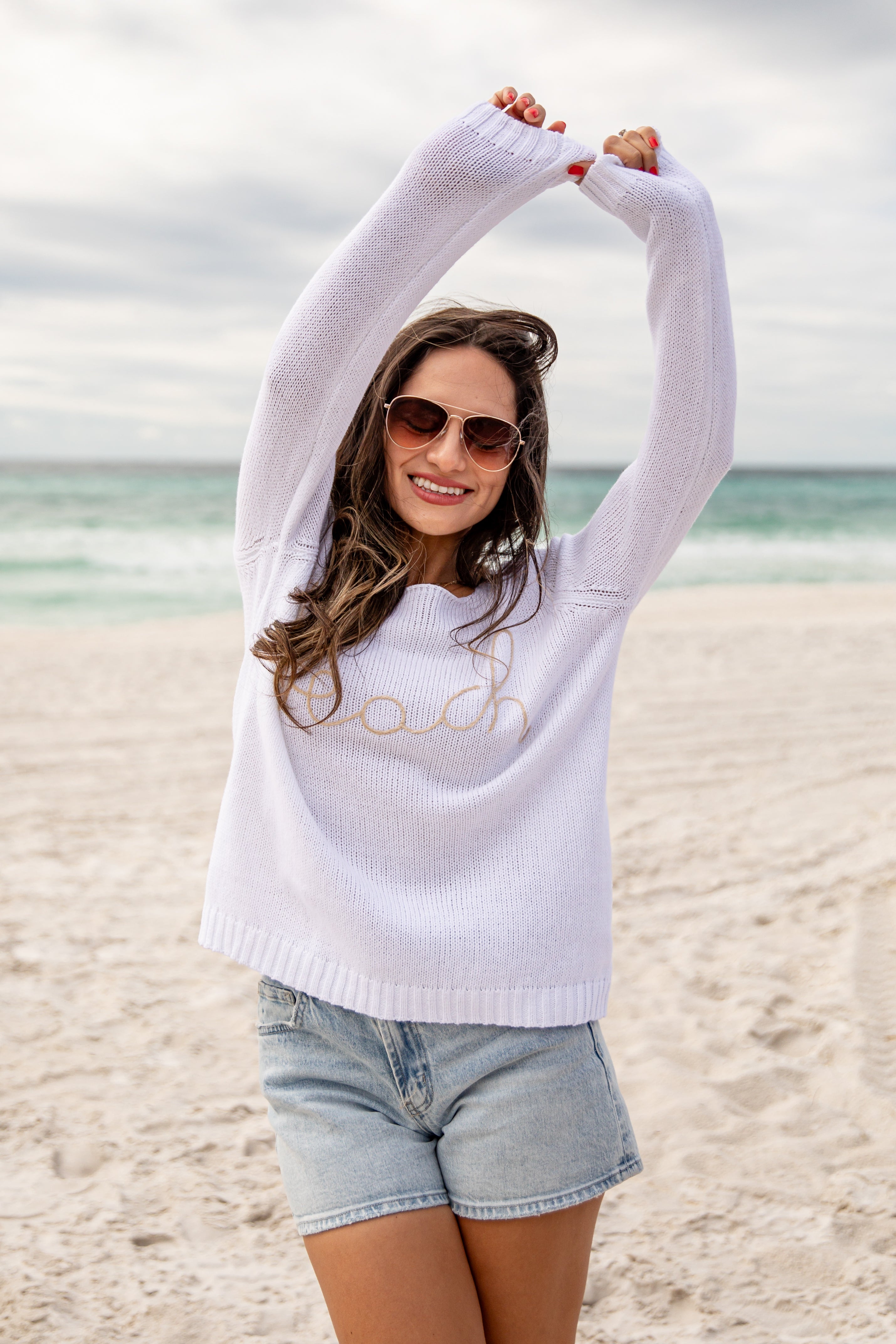 Woman on a beach wearing a white sweater with text and denim shorts, stretching her arms above her head.