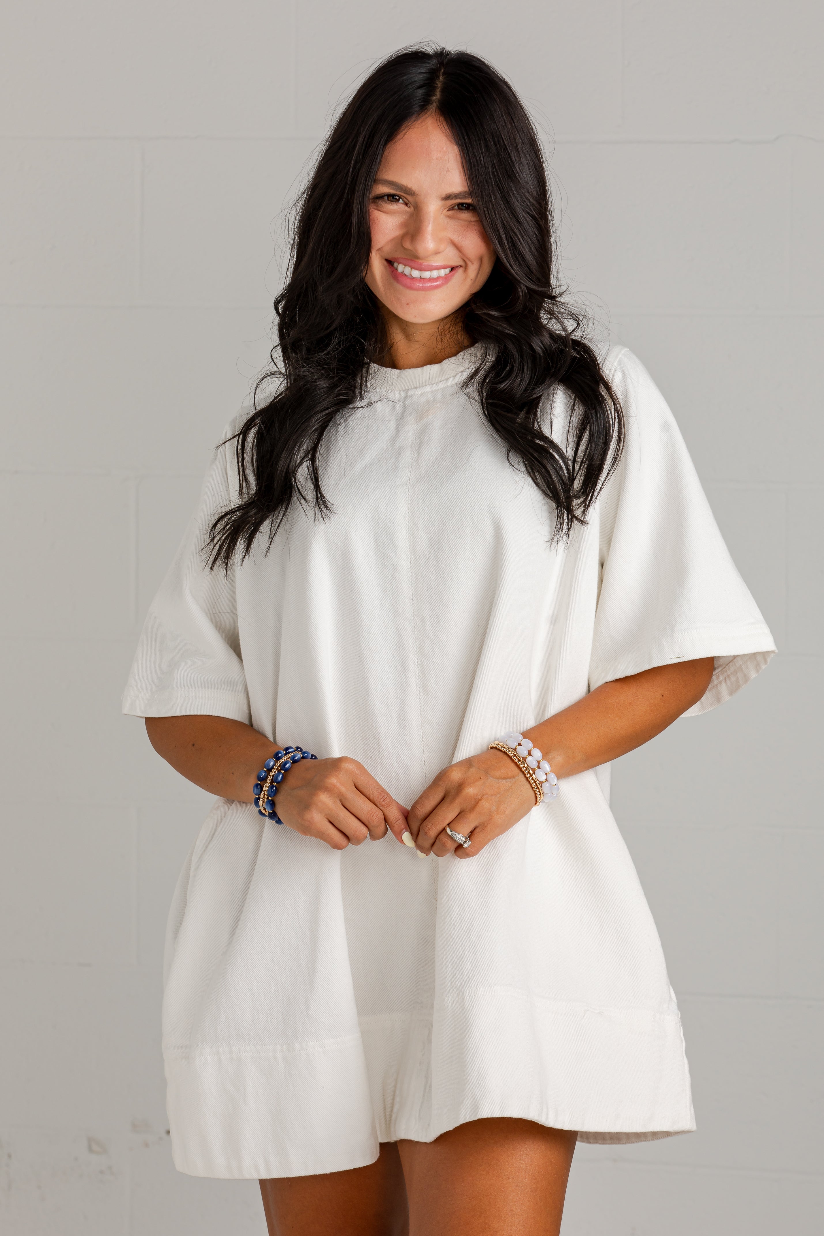 A woman with long dark hair wearing a loose-fitting, short-sleeved white linen dress or tunic with a gathered neckline, standing against a light gray brick wall.