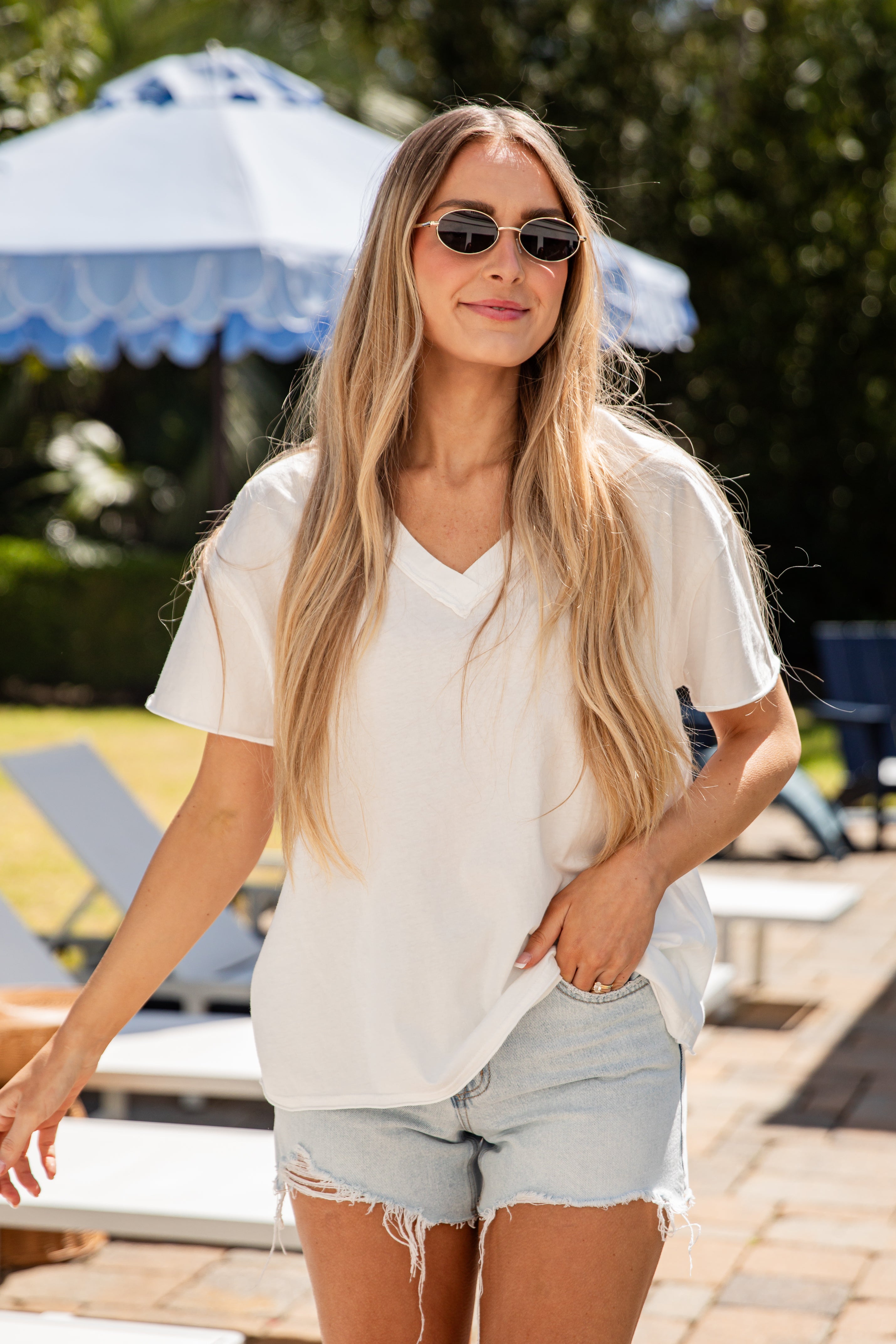 Woman wearing sunglasses and a white shirt with denim shorts in a sunlit outdoor setting.