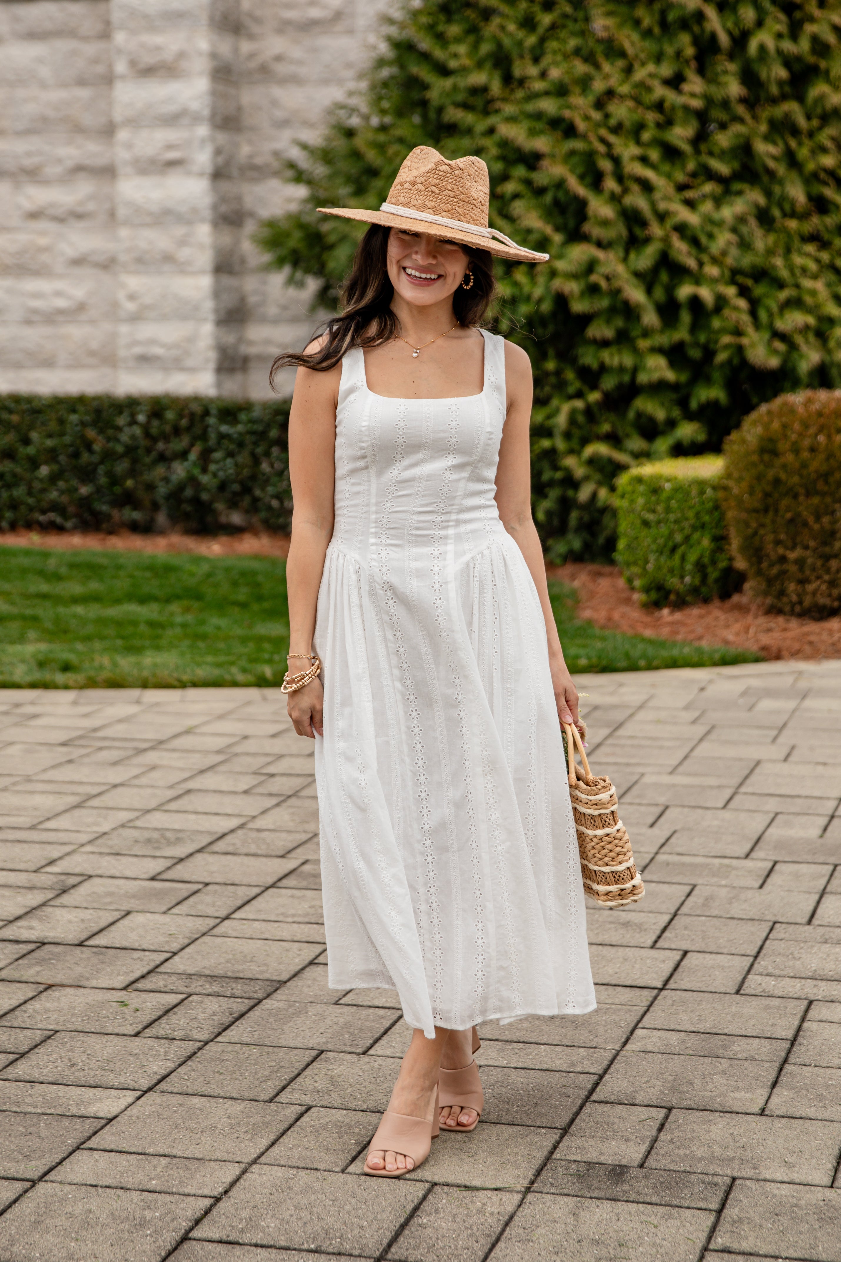 Woman in a white dress and straw hat standing on a paved walkway with greenery in the background