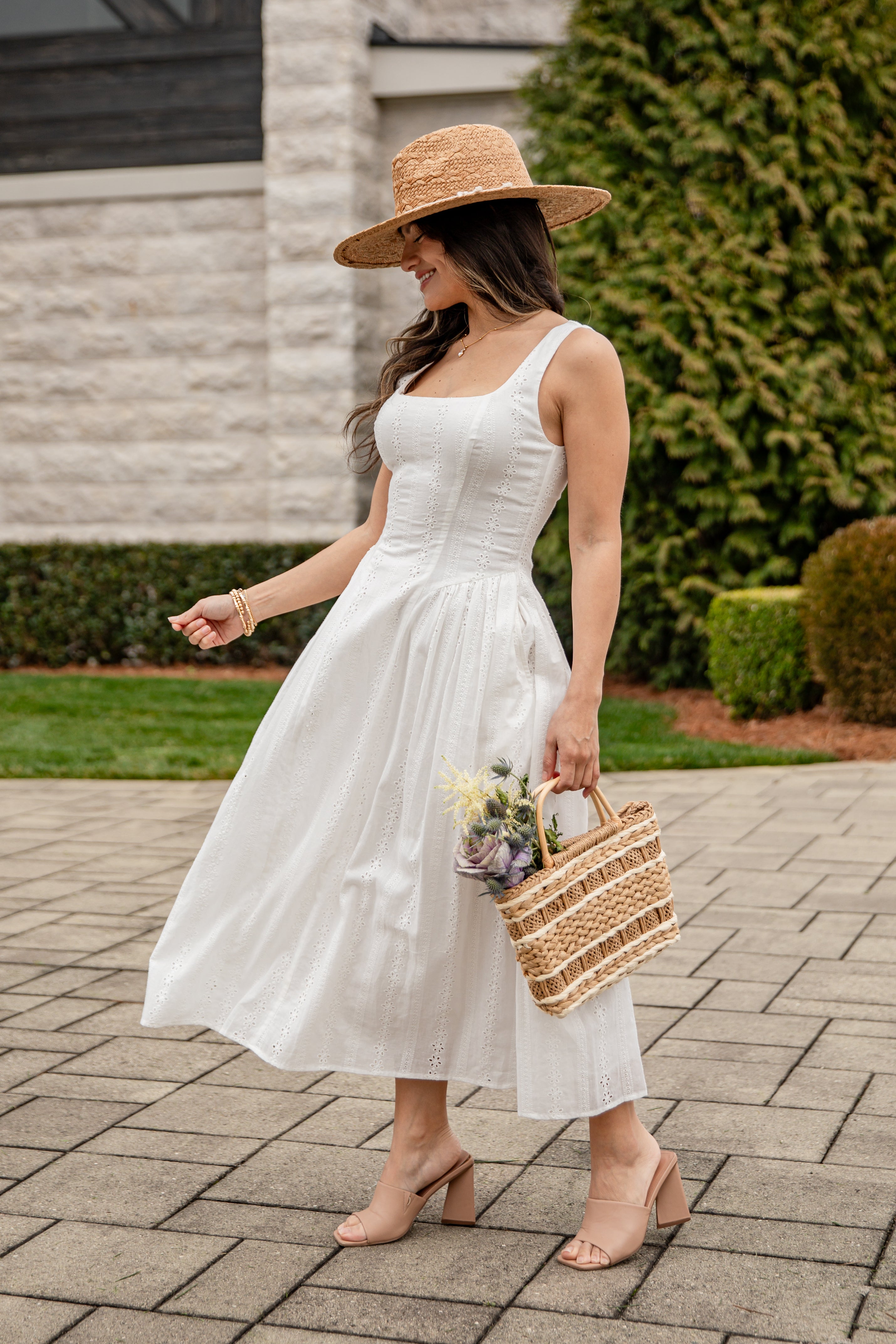 Woman in a white dress and straw hat holding a woven bag on a paved walkway.