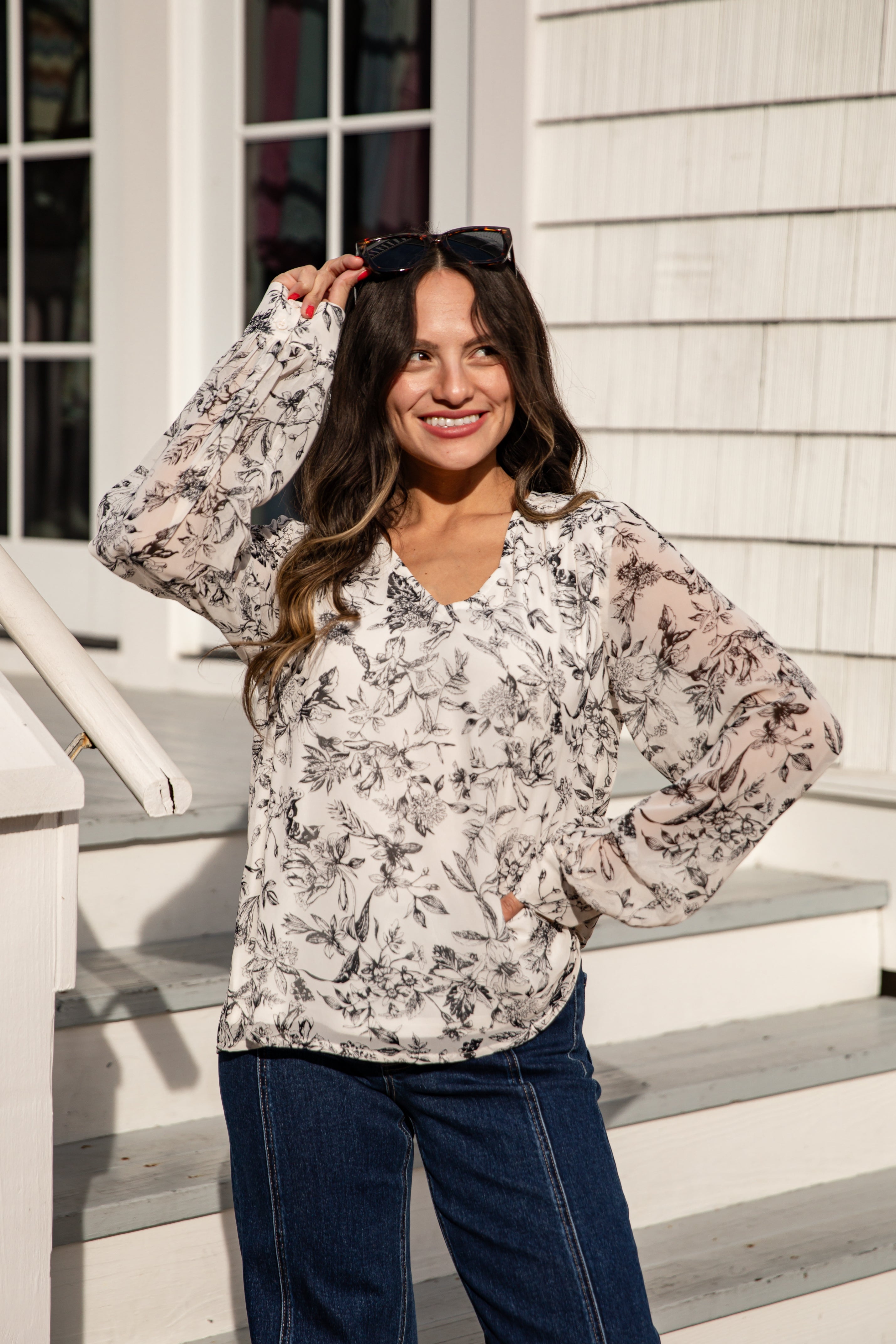 Woman wearing a floral blouse and sunglasses, standing on steps outside a house.