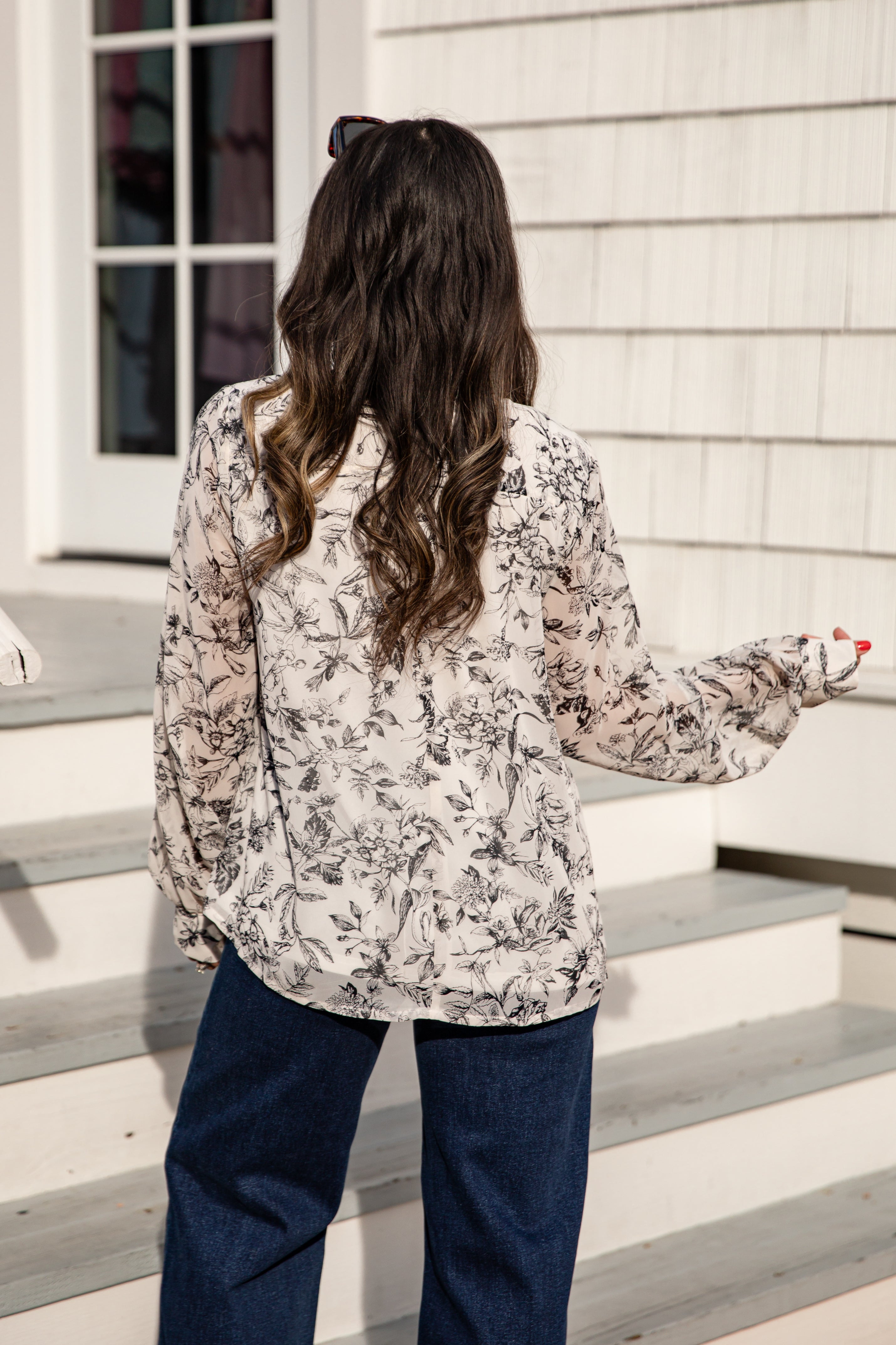 Woman standing on a set of stairs wearing a floral blouse and dark pants.