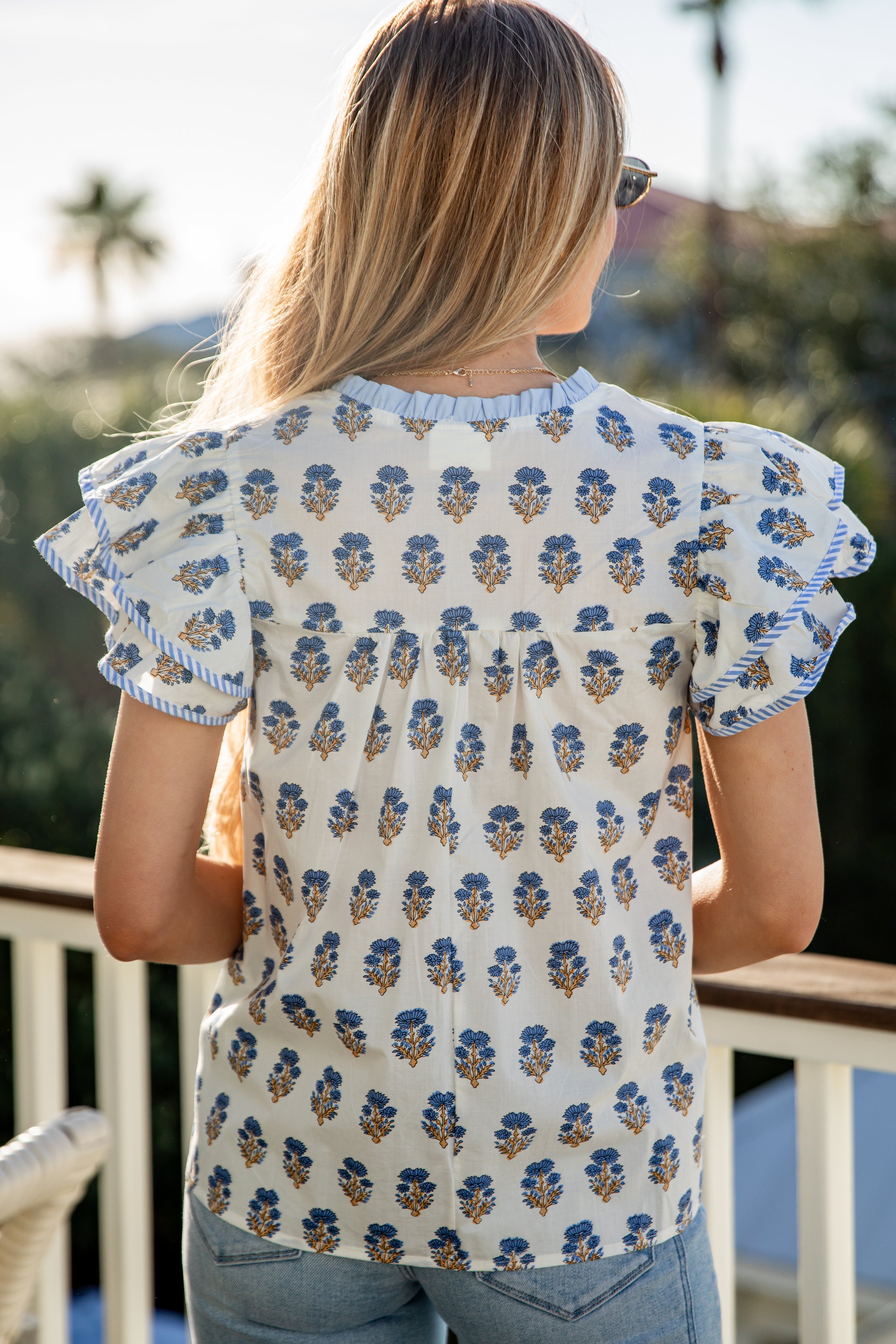 Woman wearing a floral patterned top with ruffled sleeves, standing on a balcony.
