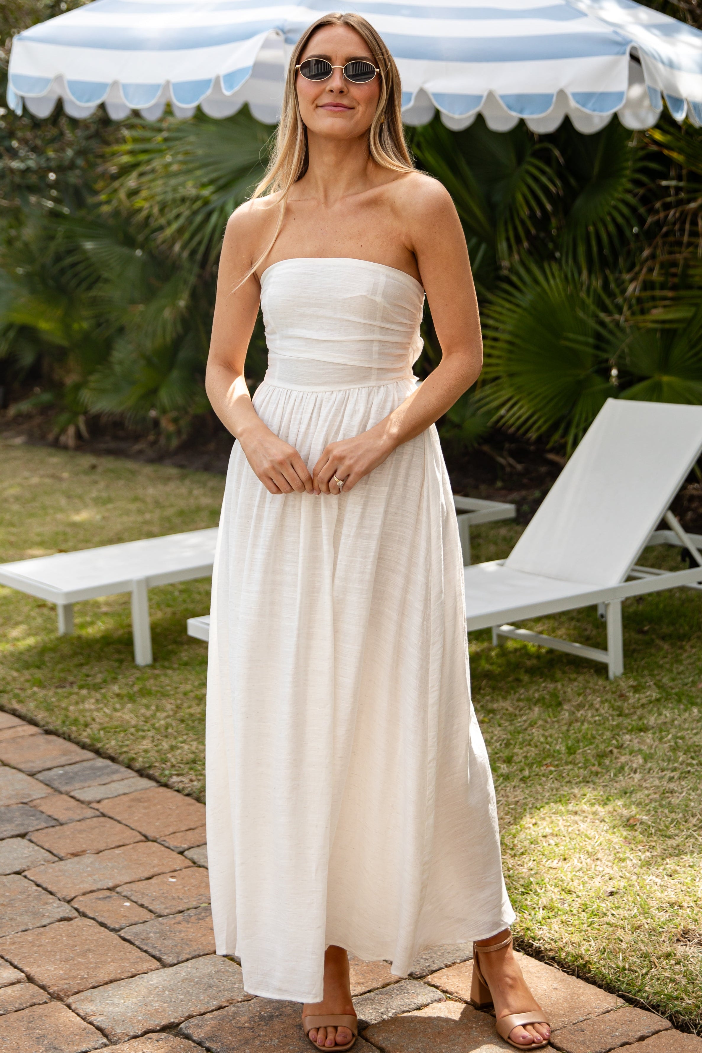 Woman in a strapless white dress standing under a striped umbrella in a garden setting.