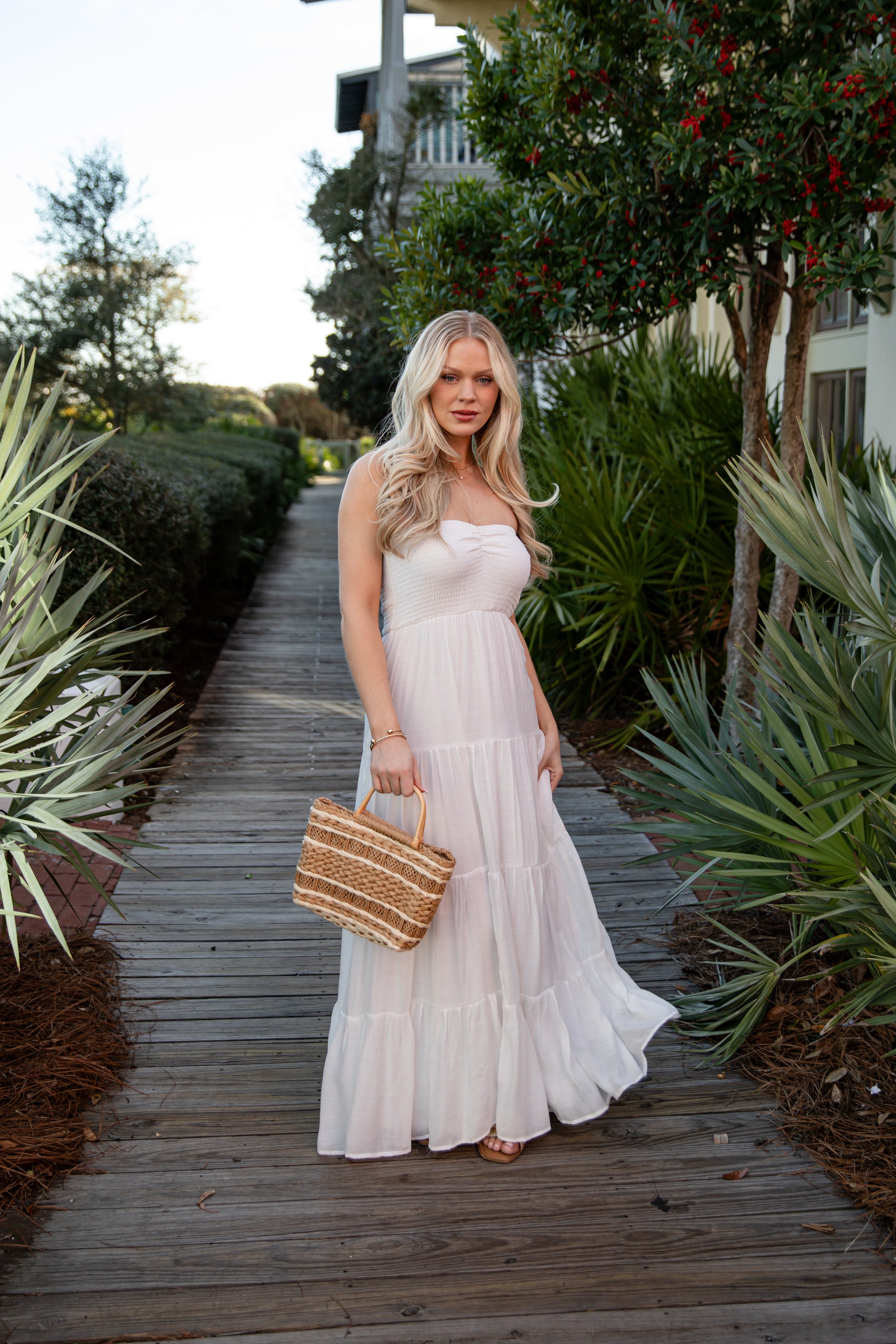 Woman in a white dress holding a straw bag on a wooden path with greenery.