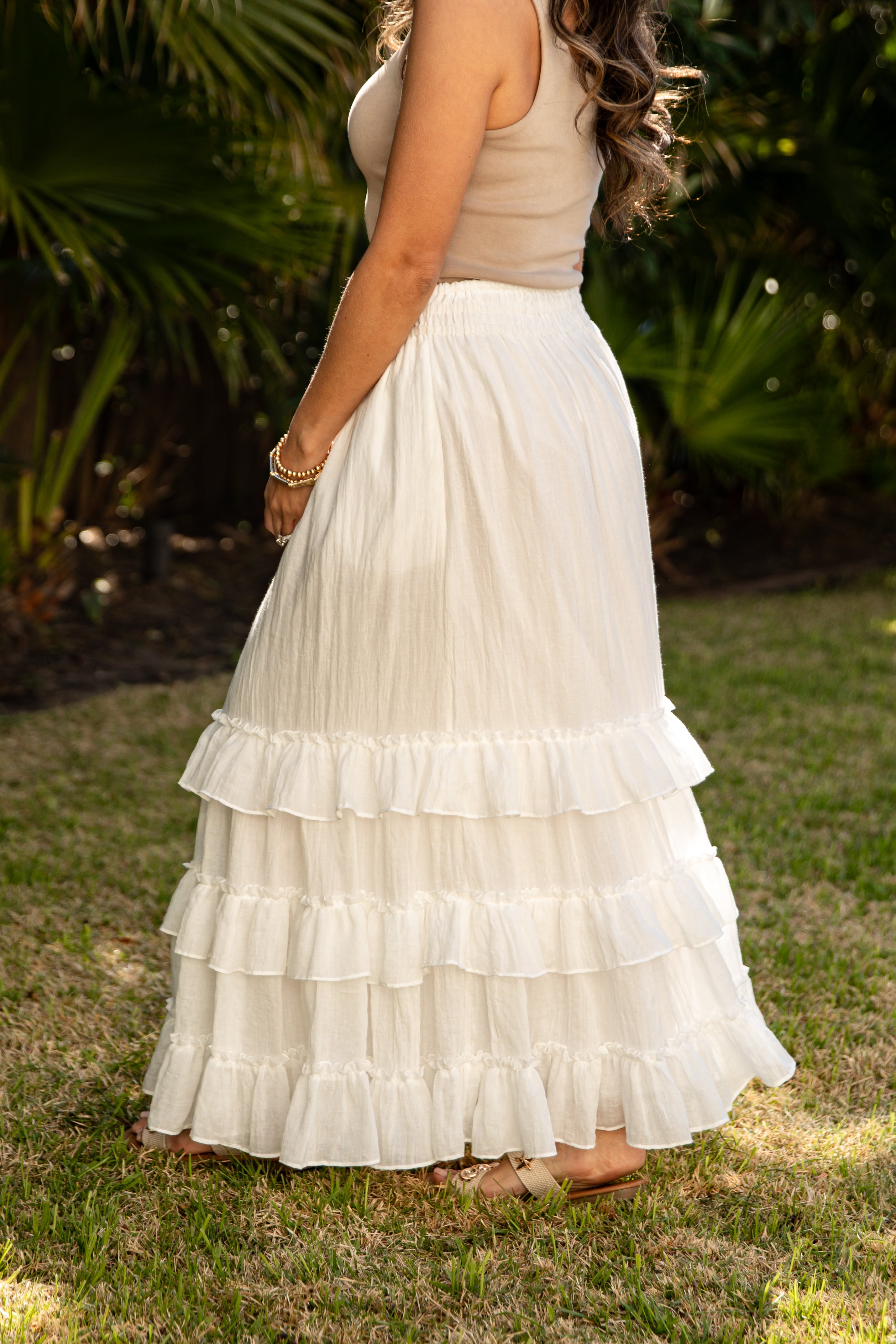Woman wearing a beige top and white tiered skirt standing on grass with greenery in the background
