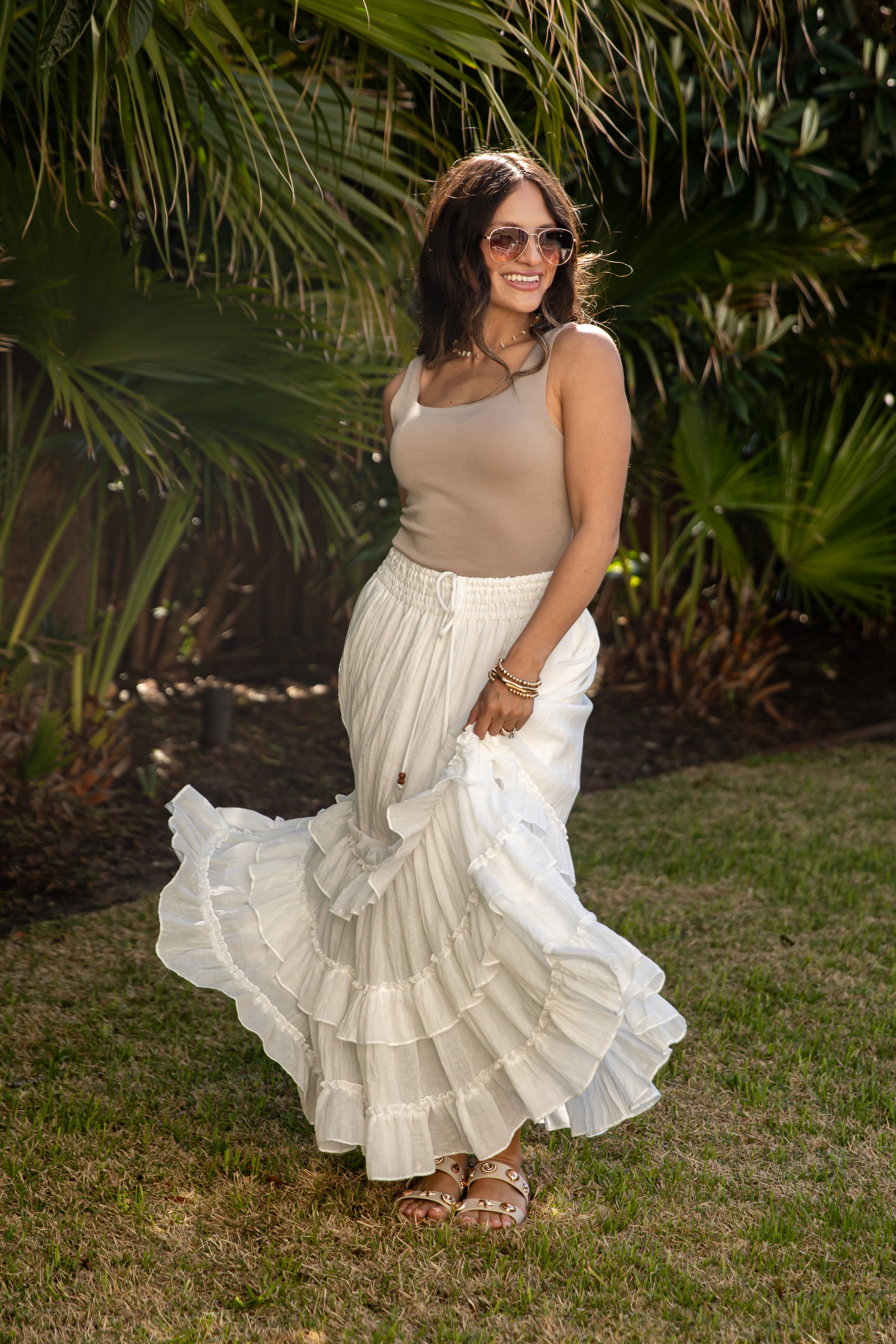 Woman in a beige top and white ruffled skirt standing outdoors with greenery in the background