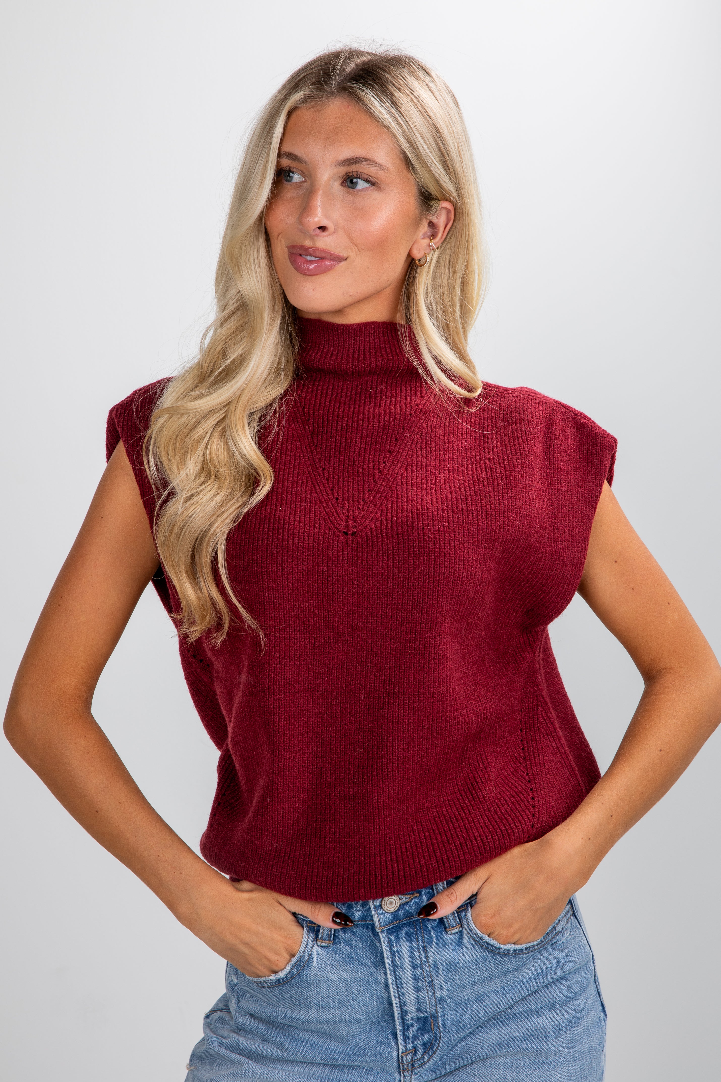 Woman wearing a burgundy sleeveless sweater and blue jeans against a white background