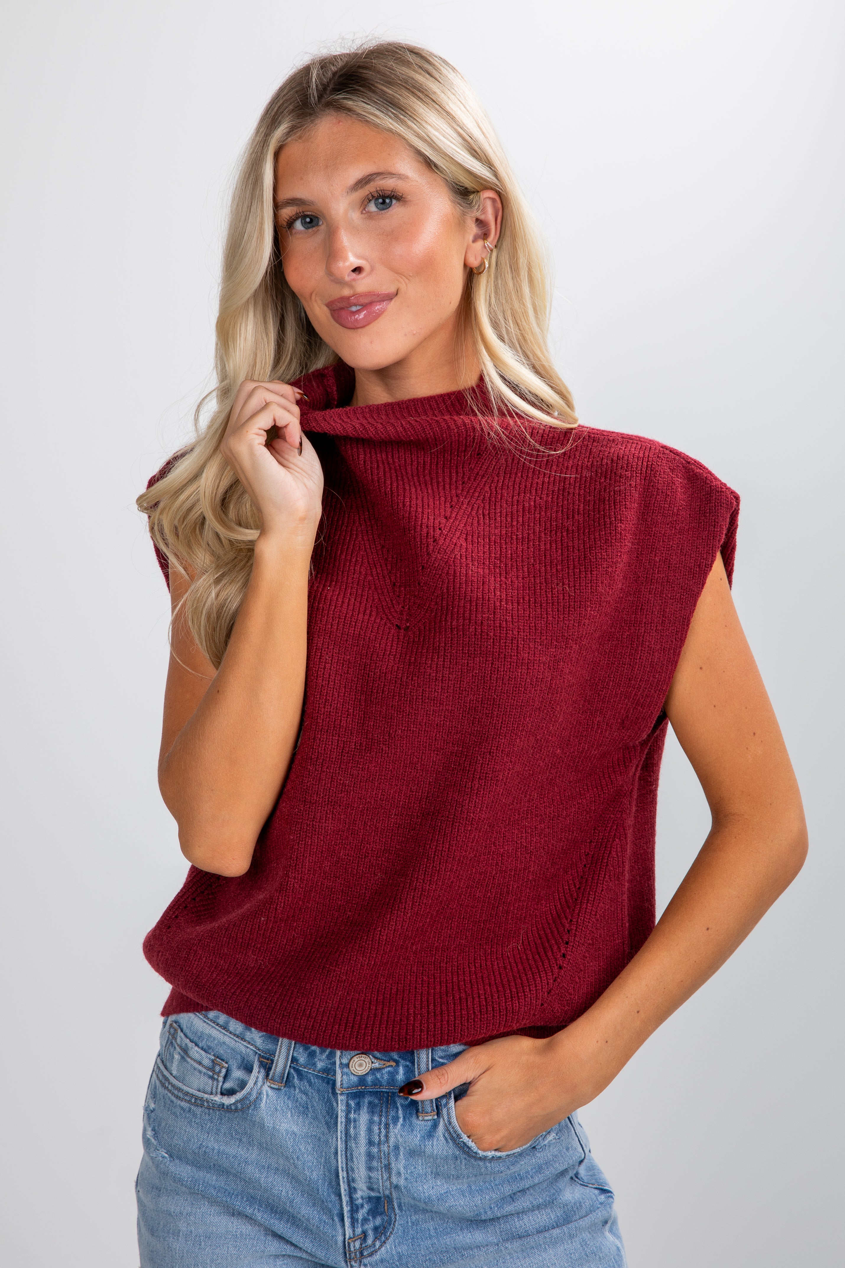 Woman wearing a red sleeveless sweater and blue jeans against a plain background