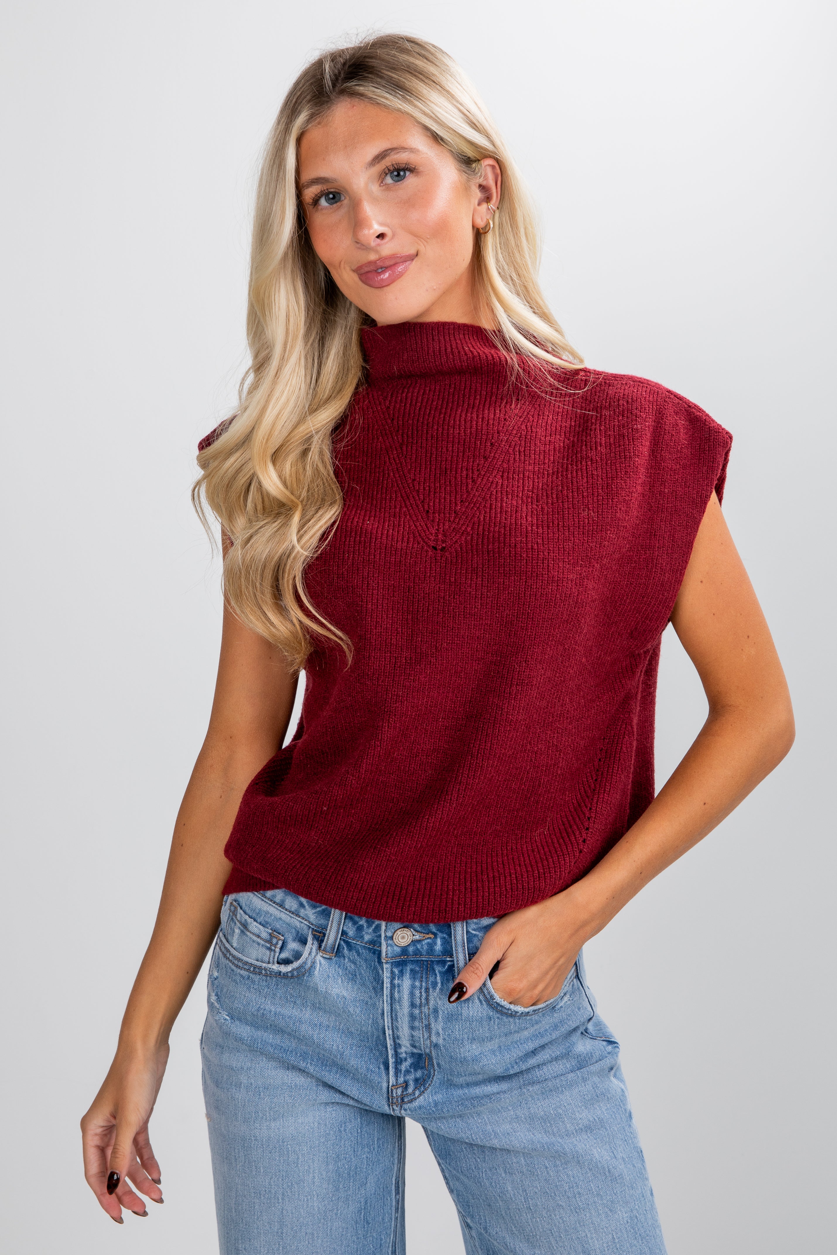 Woman wearing a red sleeveless sweater and blue jeans against a plain background