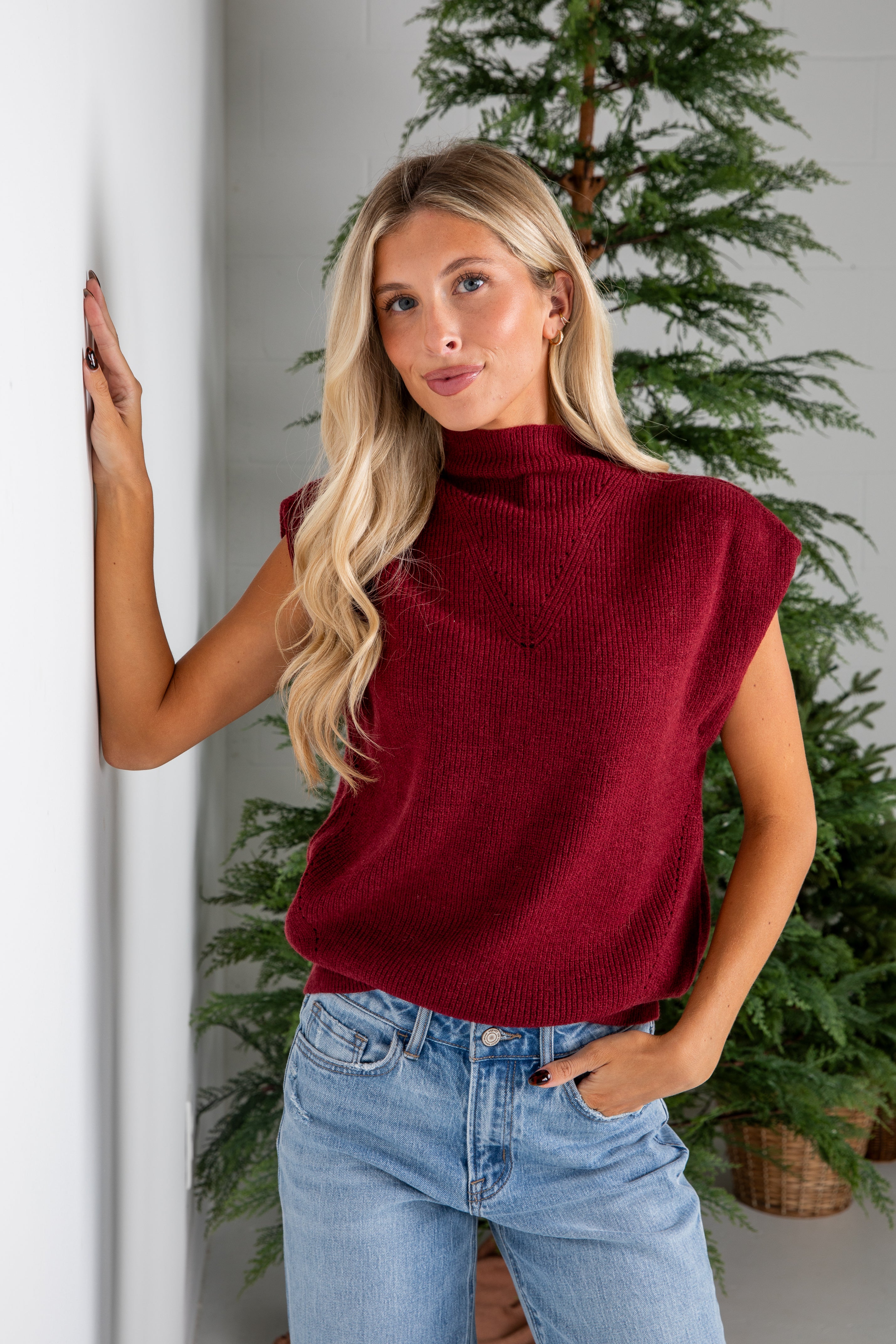 Woman wearing a red sweater and blue jeans standing against a white wall with plants in the background