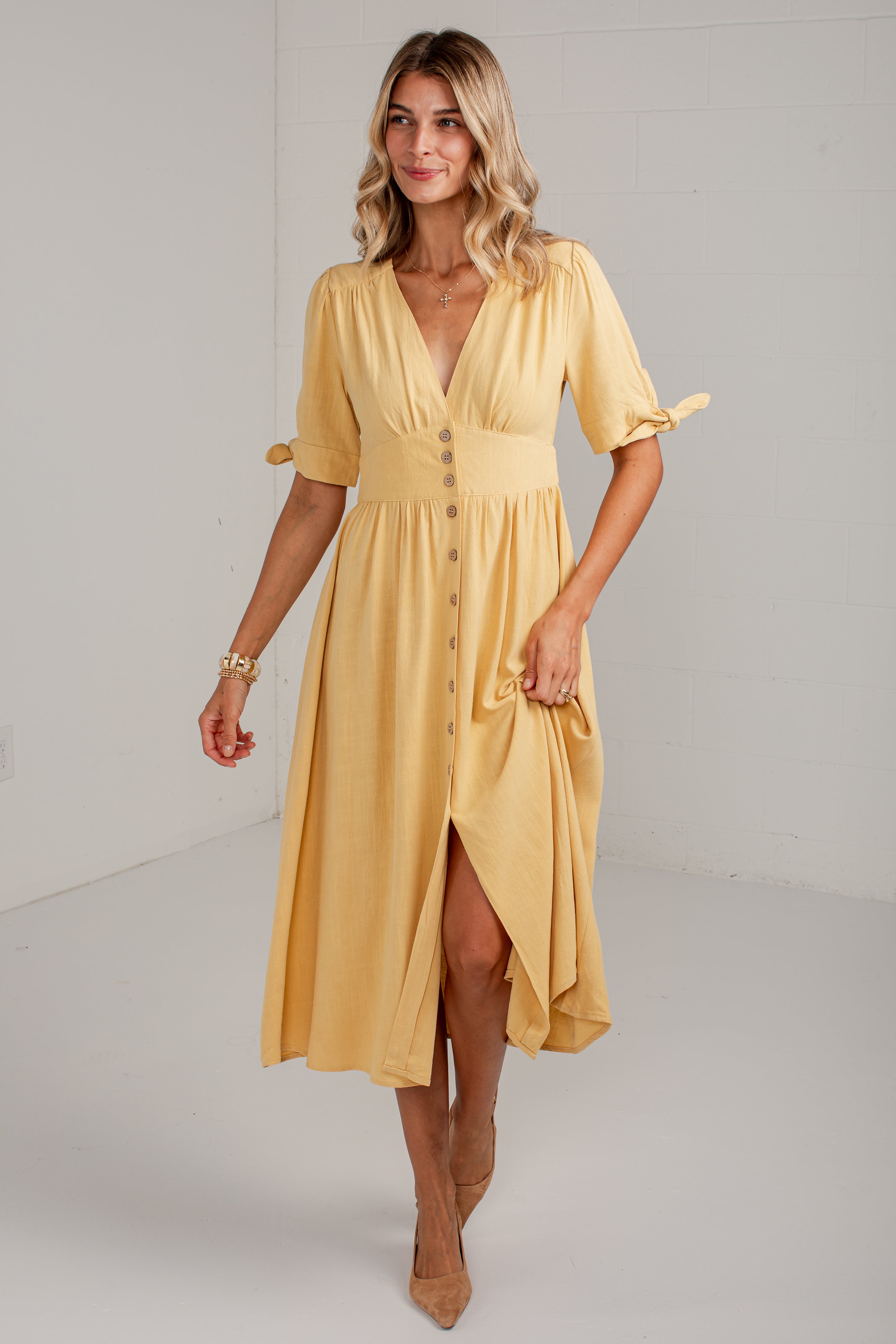 A woman modeling a mustard yellow midi dress with short flutter sleeves, a V-neckline, button-front detail, and a high-low hem, photographed in a bright white studio setting.