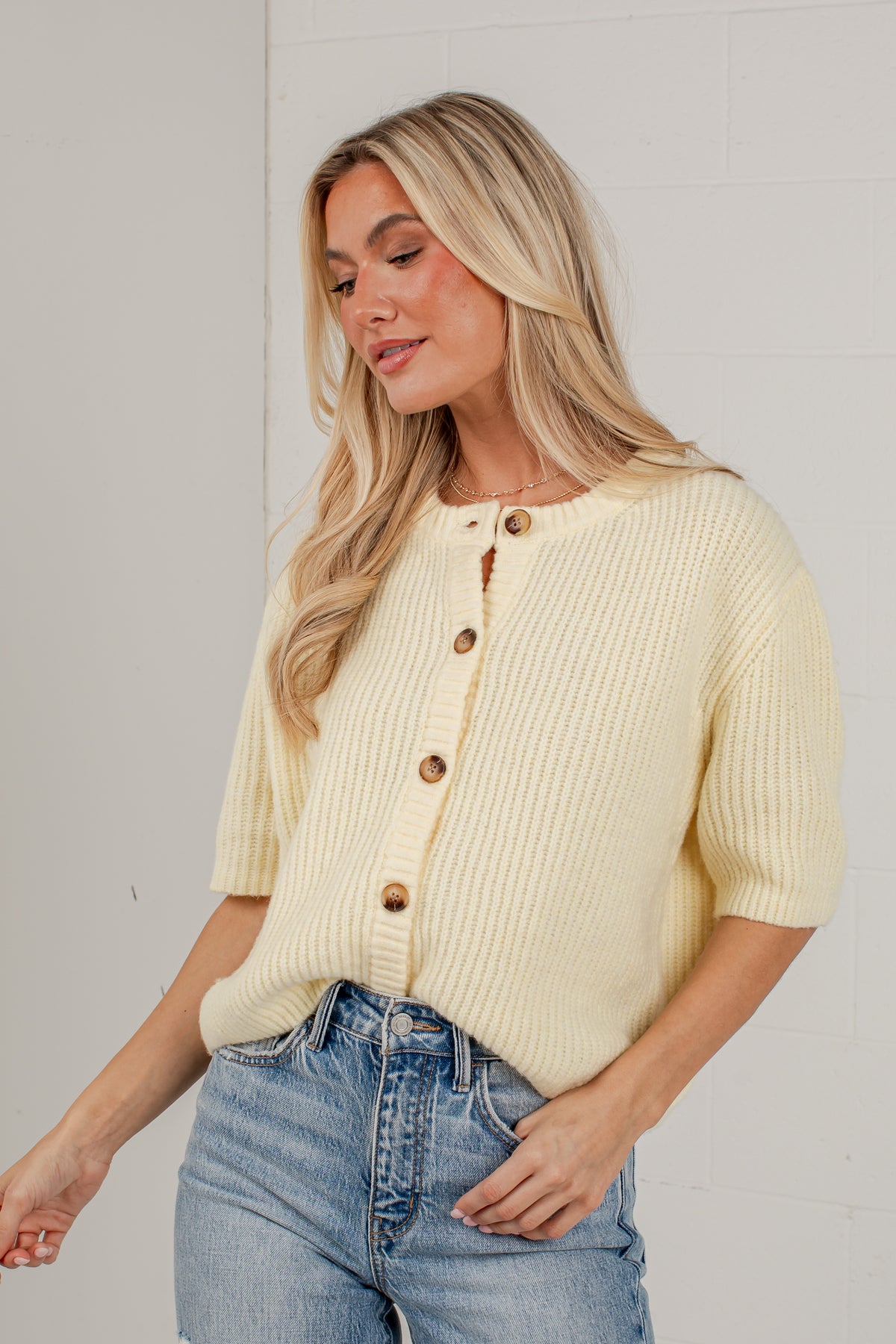 A woman wearing a pale yellow ribbed knit short-sleeve cardigan with brown buttons, paired with light-wash blue jeans, standing against a white brick wall.