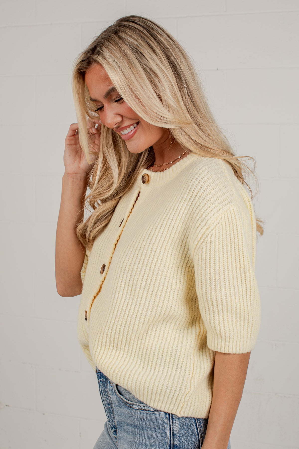 A woman with blonde hair wearing a pale yellow ribbed knit cardigan with brown buttons, paired with blue jeans, posing against a white brick wall.