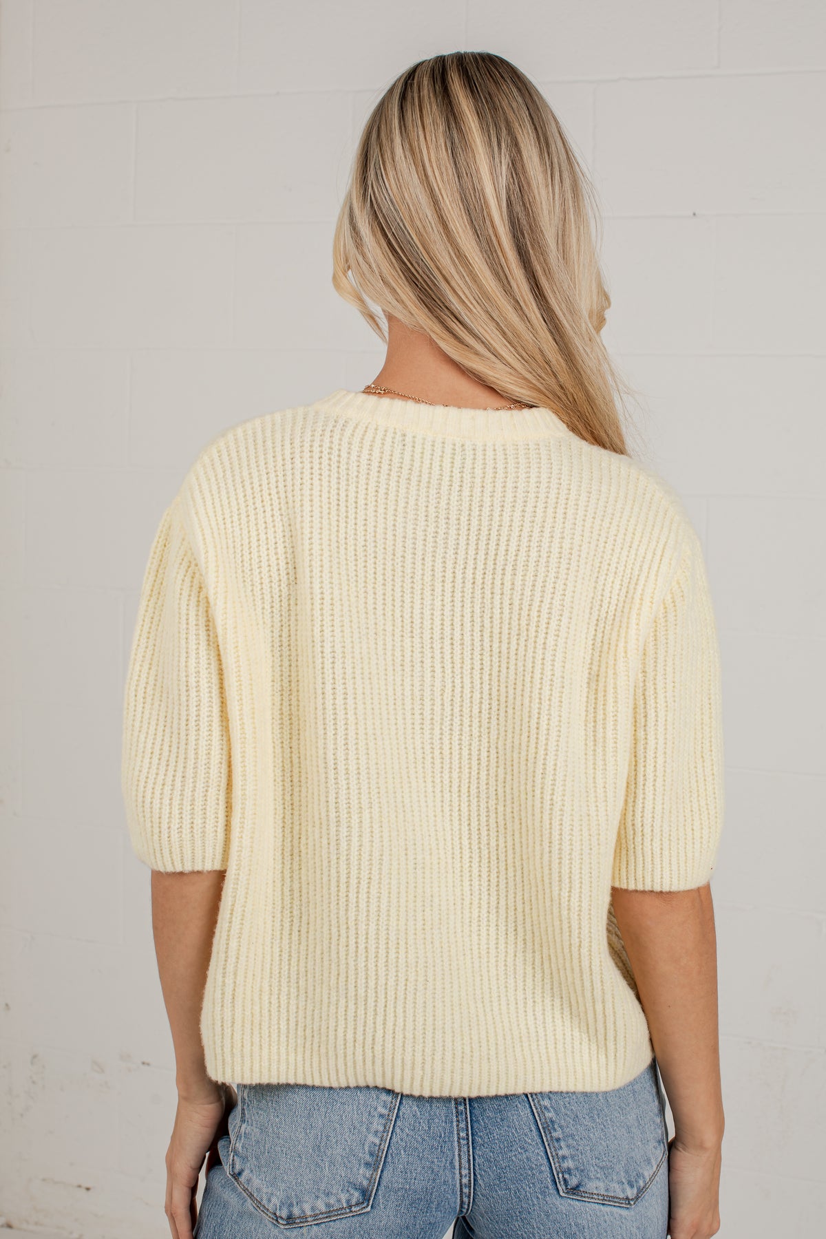 A woman viewed from behind wearing a cream-colored ribbed knit sweater and light blue jeans, standing against a white brick wall.