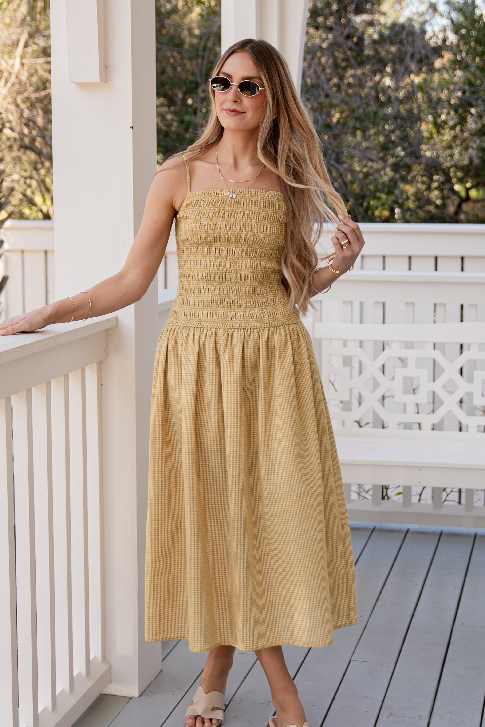 Woman in a yellow dress standing on a porch with white railings and greenery in the background.
