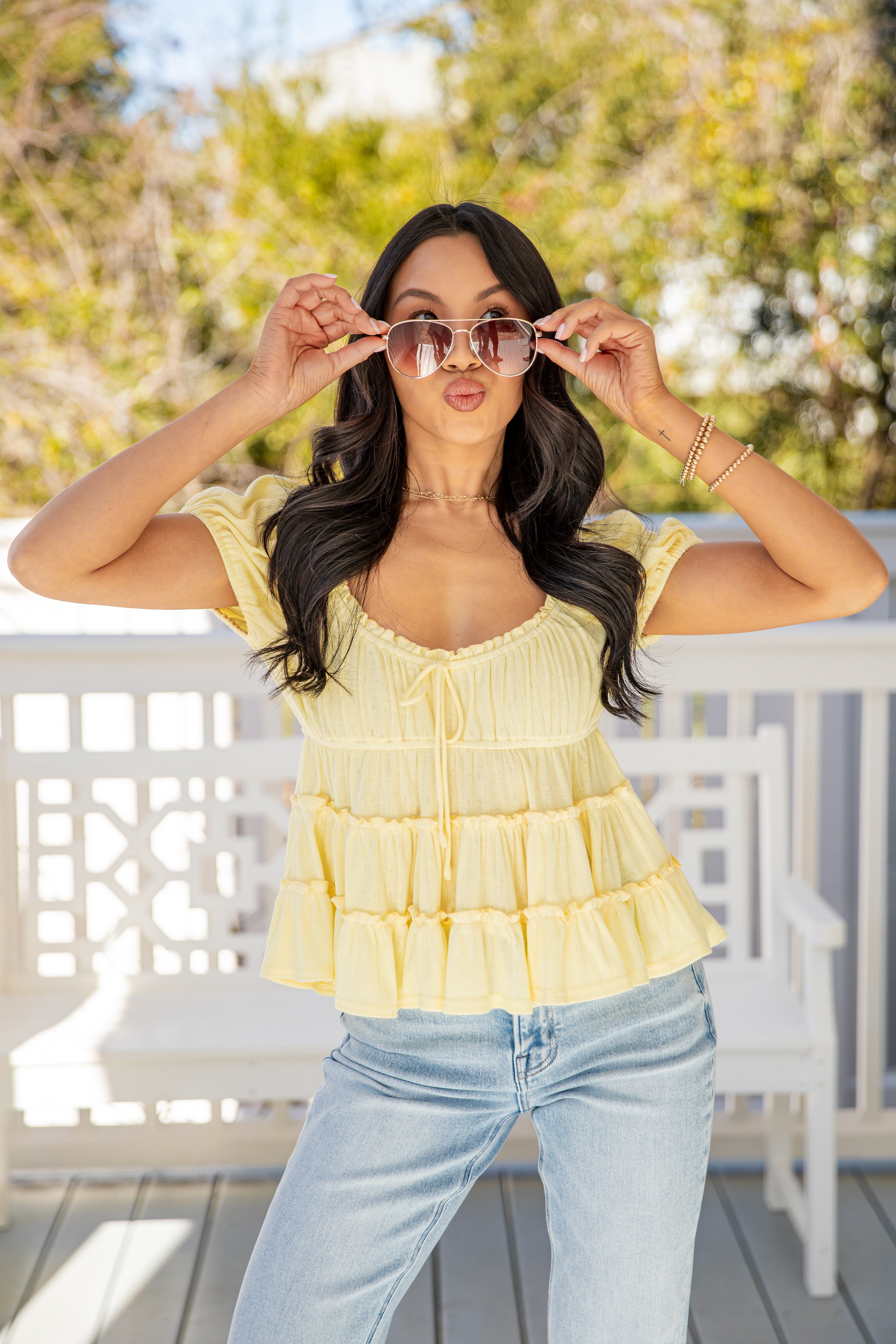 Woman wearing a yellow tiered top and light blue jeans, holding sunglasses up to her face on a sunny day.
