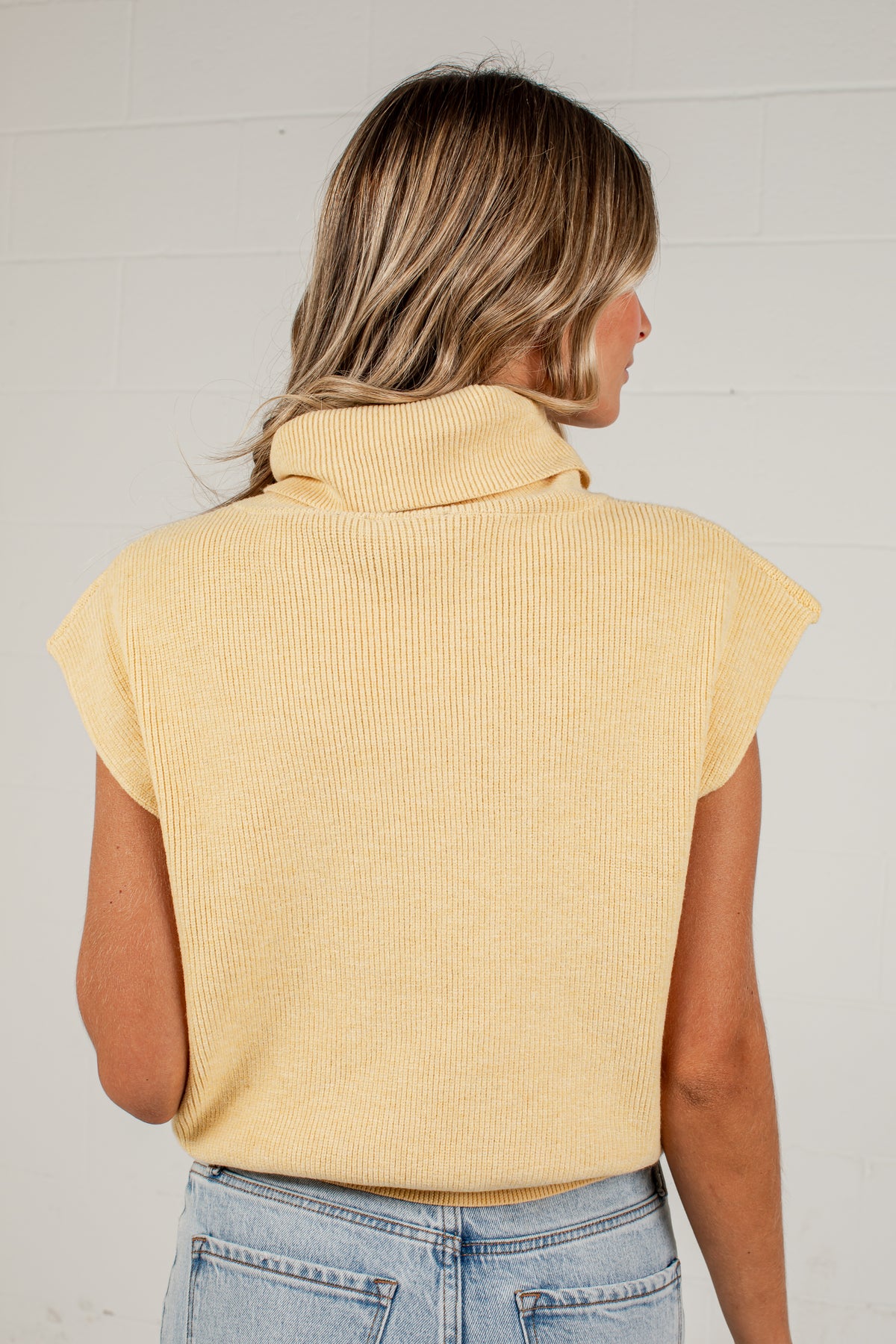 A woman viewed from the back wearing a pale yellow ribbed knit turtleneck sweater with cap sleeves and light blue jeans, standing against a white brick wall.