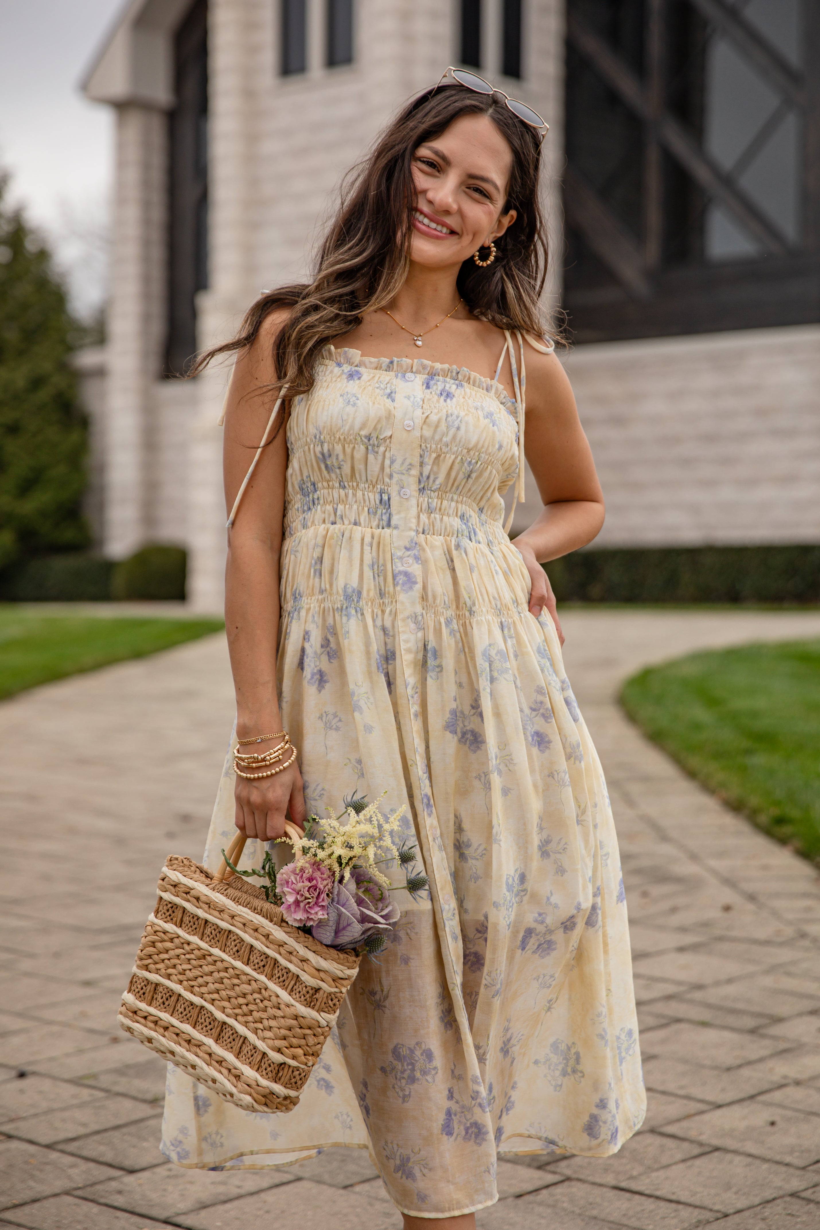 Woman in a floral dress holding a woven bag with flowers, standing on a sidewalk.