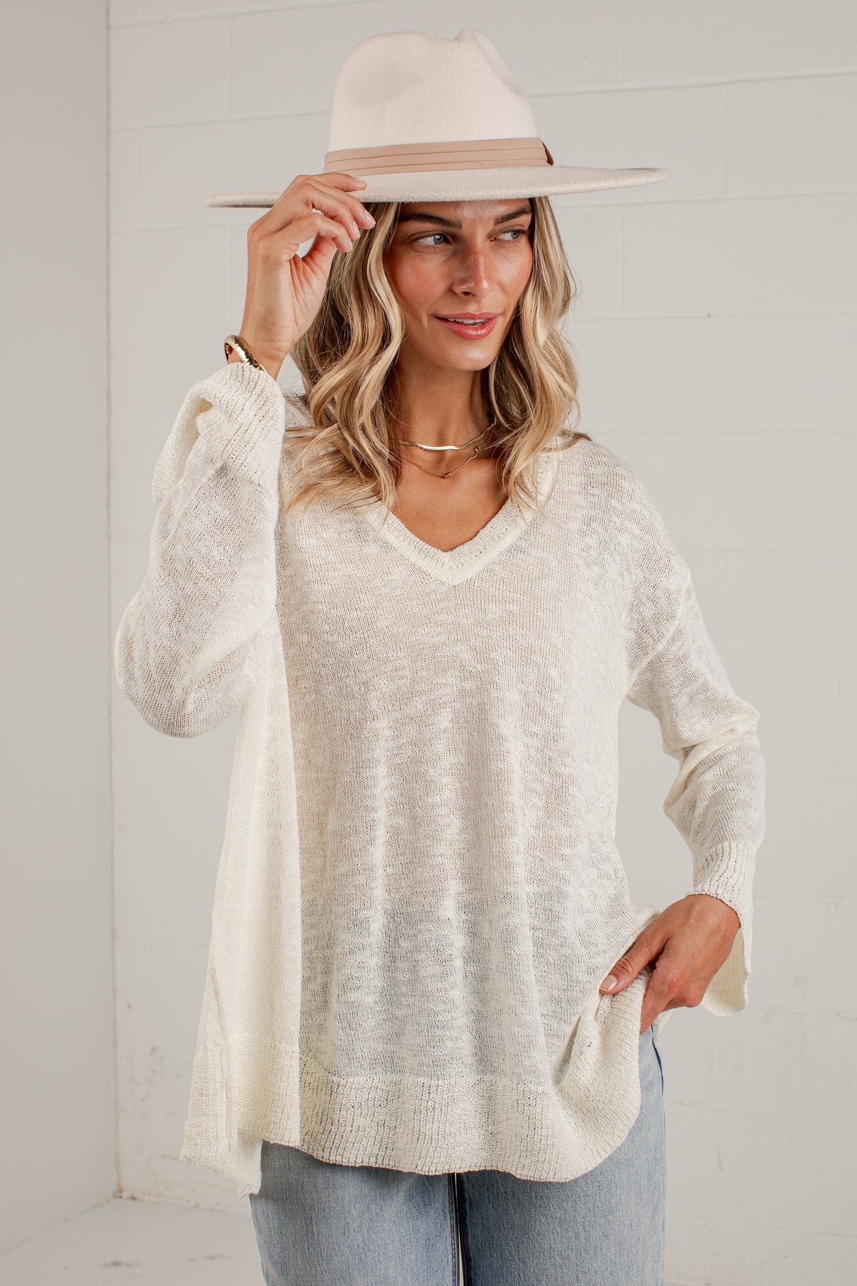 A woman wearing a cream-colored, long-sleeve knit V-neck sweater, light blue jeans, and a beige wide-brim hat, posing against a white paneled wall.