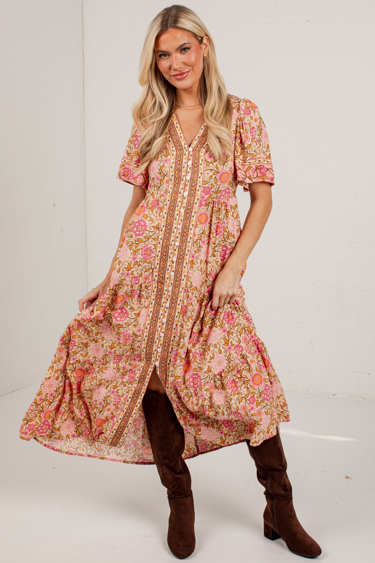 A woman modeling a bohemian-style midi dress with a pink and orange floral pattern, brown buttons, and puff sleeves, paired with brown suede boots against a white studio background.