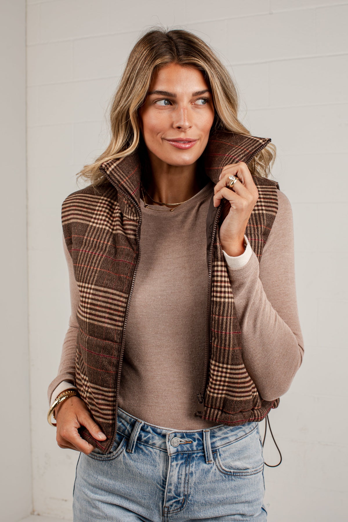 A woman wearing a brown and beige plaid zip-up puffer vest over a taupe ribbed long-sleeve top and light blue jeans, standing against a white paneled wall.