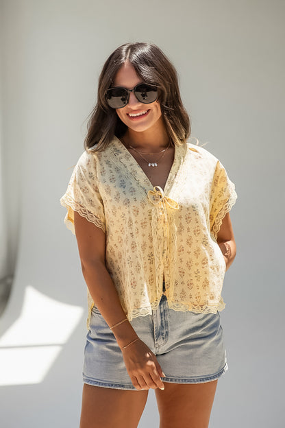 A woman wearing a light yellow floral short-sleeve tie-front blouse with lace trim and denim shorts, standing against a plain light gray wall.
