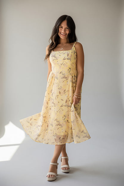 A woman modeling a light yellow sleeveless midi dress with a delicate leaf pattern, standing against a neutral light gray studio background.