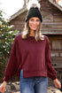 Woman wearing a burgundy sweater and black beanie standing in front of a wooden cabin.