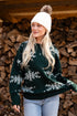 Woman wearing a green sweater with snowflake patterns and a white beanie in front of stacked firewood.