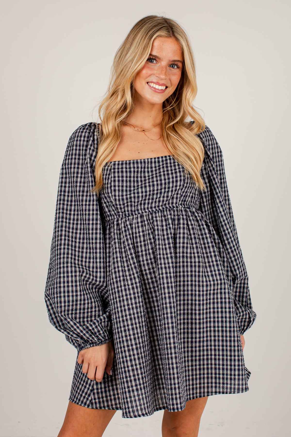 A woman modeling a navy and white gingham mini dress with a square neckline, smocked bodice, and voluminous long puff sleeves against a plain light gray studio background.