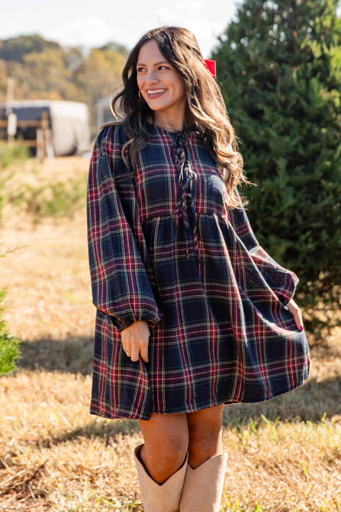 Woman in a plaid dress and beige boots standing in a field with trees in the background