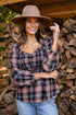 Woman wearing a plaid shirt and wide-brimmed hat standing in front of stacked wood.