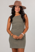 A woman modeling a sleeveless, bodycon mini dress in a muted sage green color, paired with a brown wide-brim hat, against a plain light gray studio background.