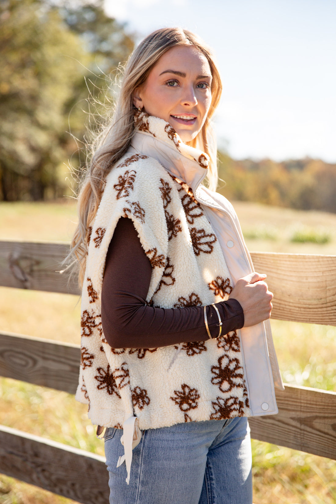 Woman wearing a white floral-patterned vest outdoors with a wooden fence and trees in the background.