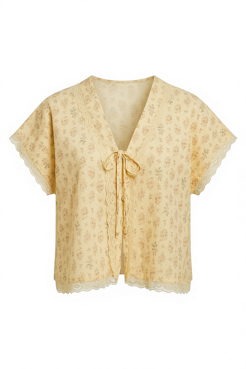 A pale yellow short-sleeve blouse with a delicate floral pattern, a V-neckline, and a front tie detail, finished with scalloped lace trim.