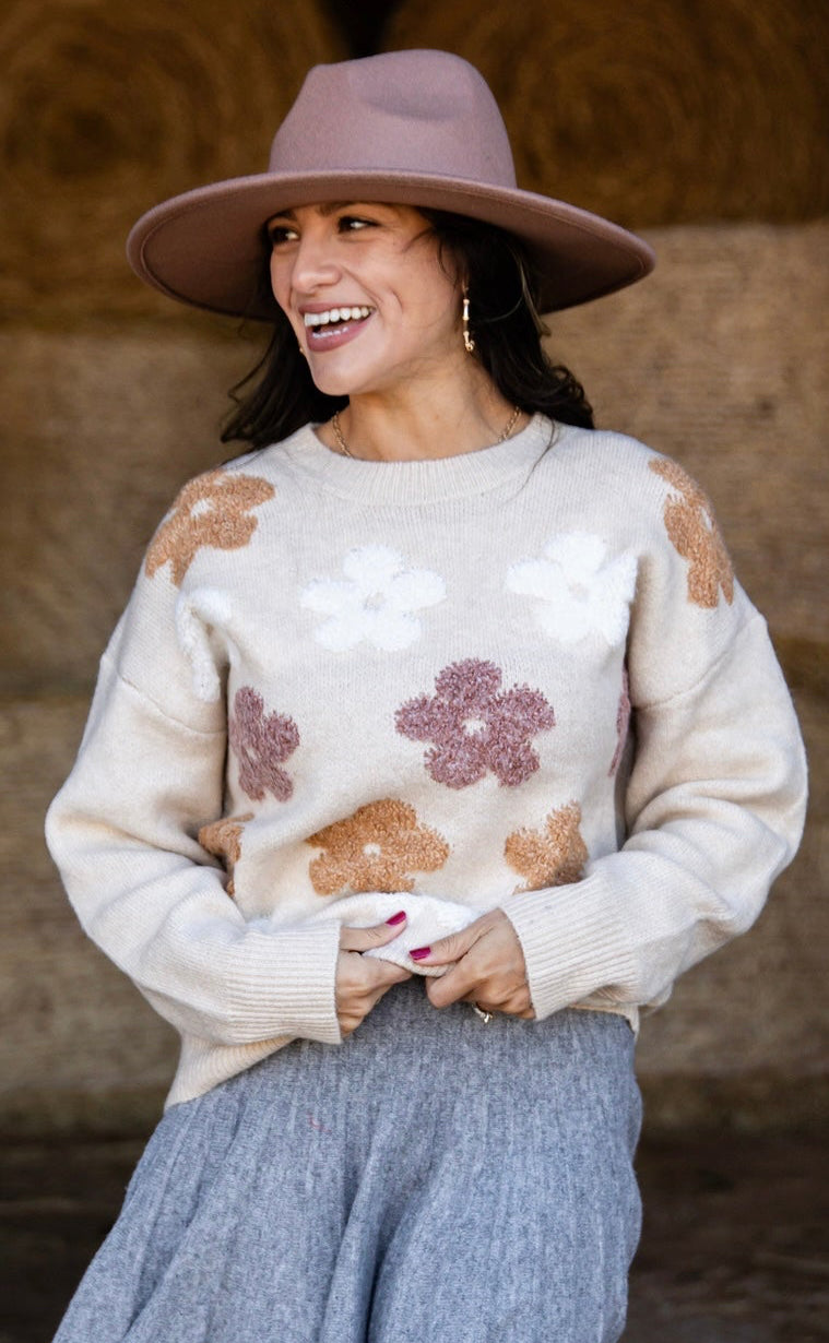 Woman wearing a patterned sweater, skirt, and cowboy boots in front of hay bales.
