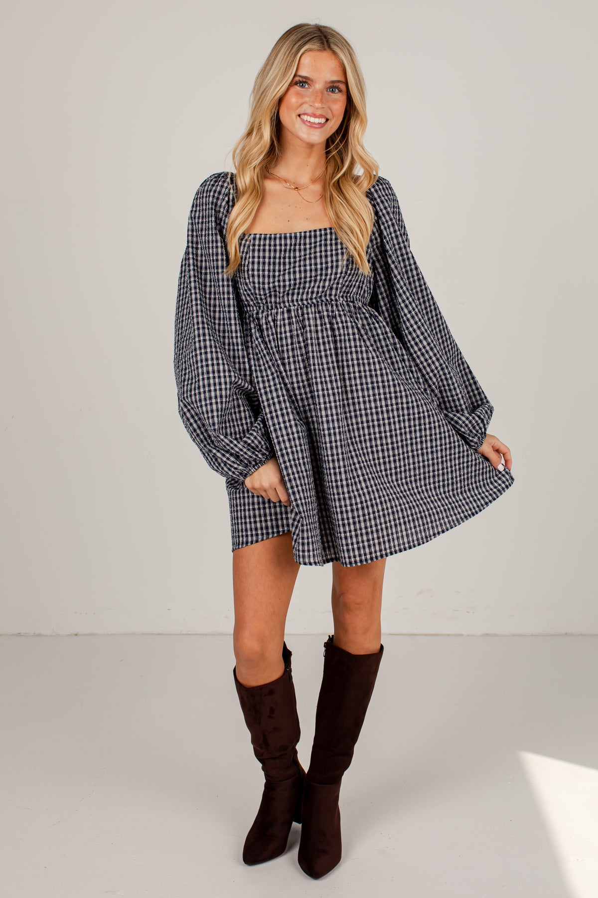 A woman modeling a black and white gingham mini dress with long balloon sleeves and a smocked bodice, paired with dark brown knee-high boots, against a plain light gray studio background.