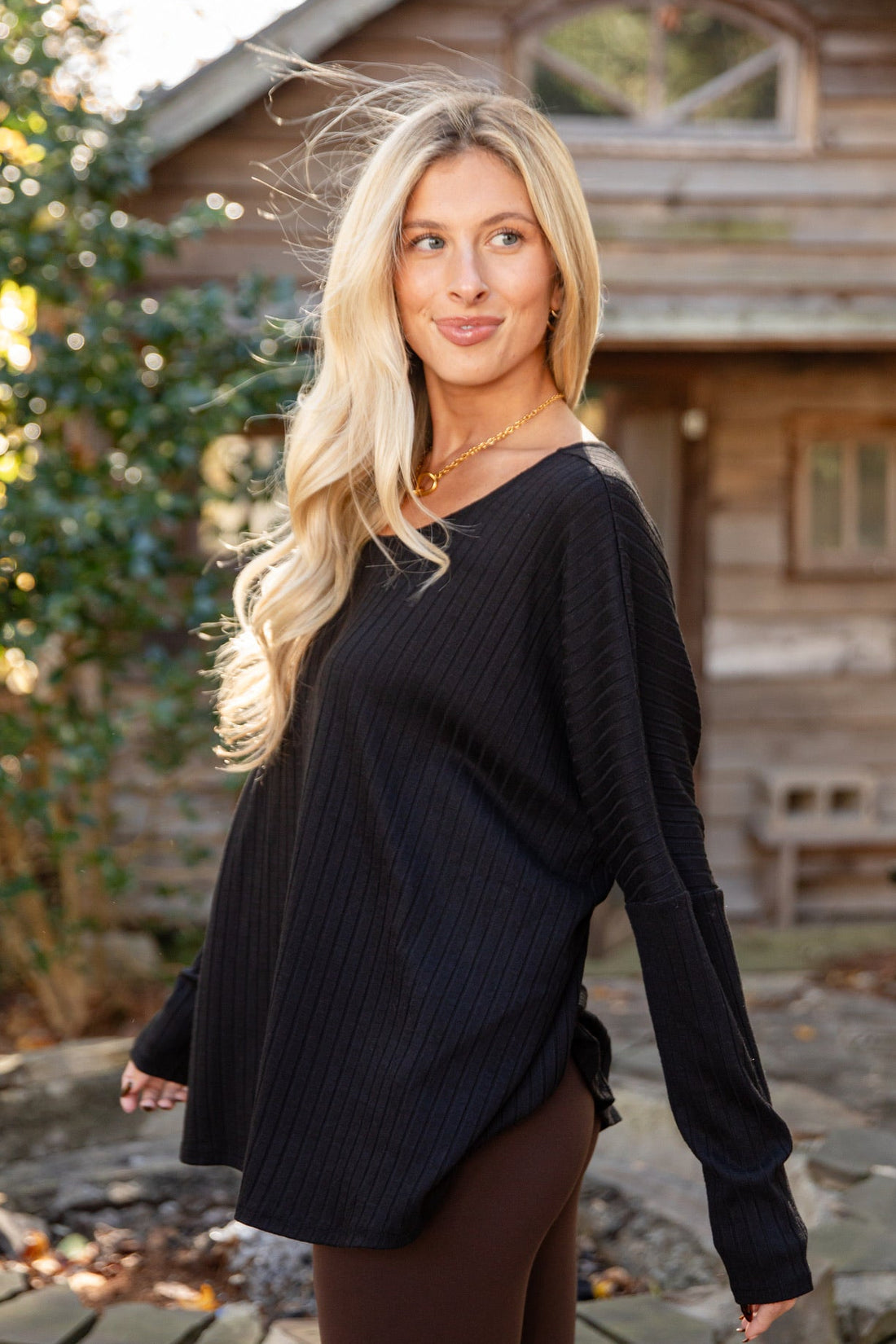Woman in a black sweater and brown pants standing outdoors with a wooden cabin in the background.
