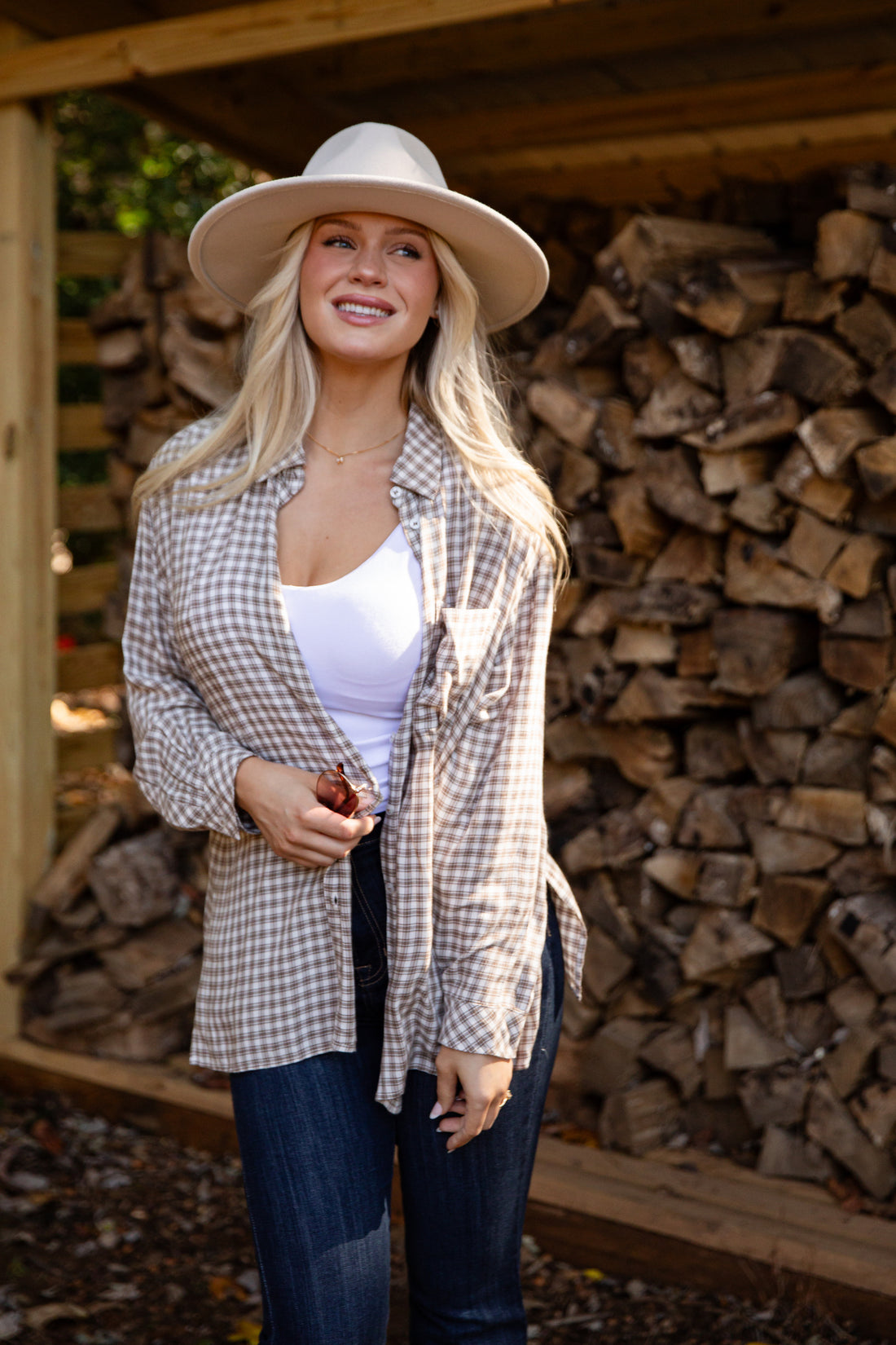 Woman wearing a plaid shirt, white top, and wide-brimmed hat standing in front of stacked firewood.