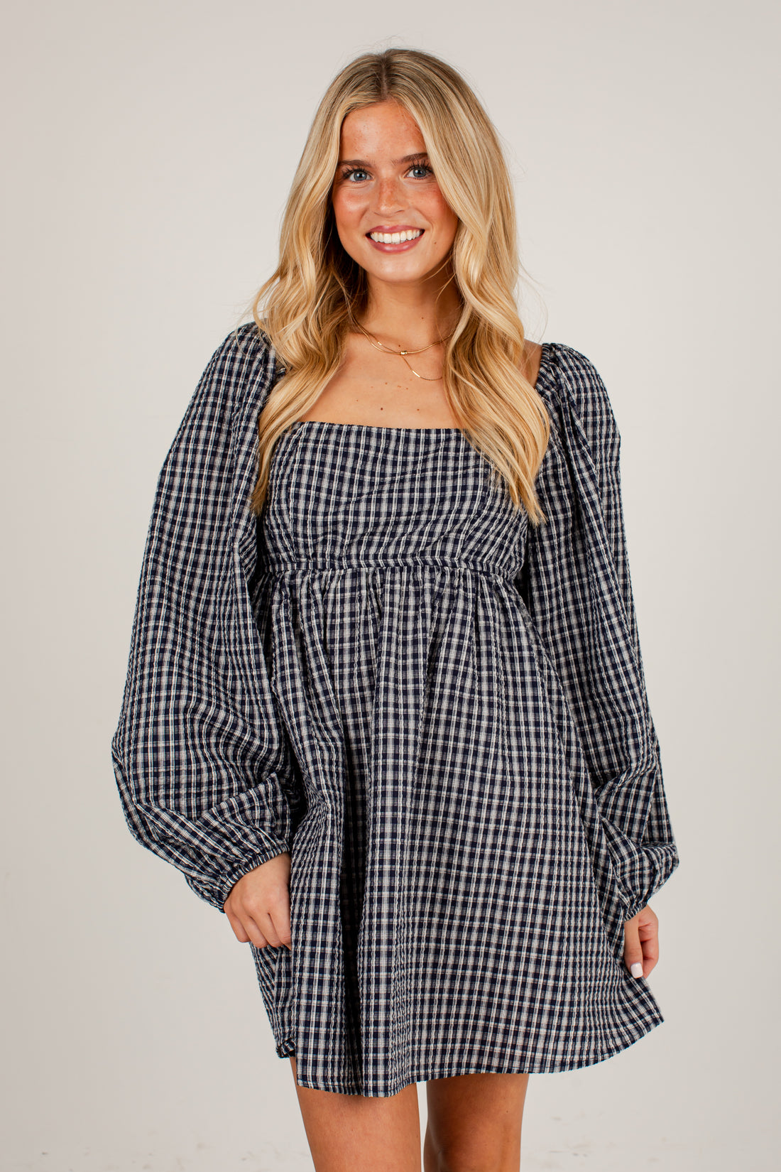 A woman modeling a black and white gingham mini dress with a square neckline, smocked waist, and voluminous balloon sleeves against a plain light beige background.