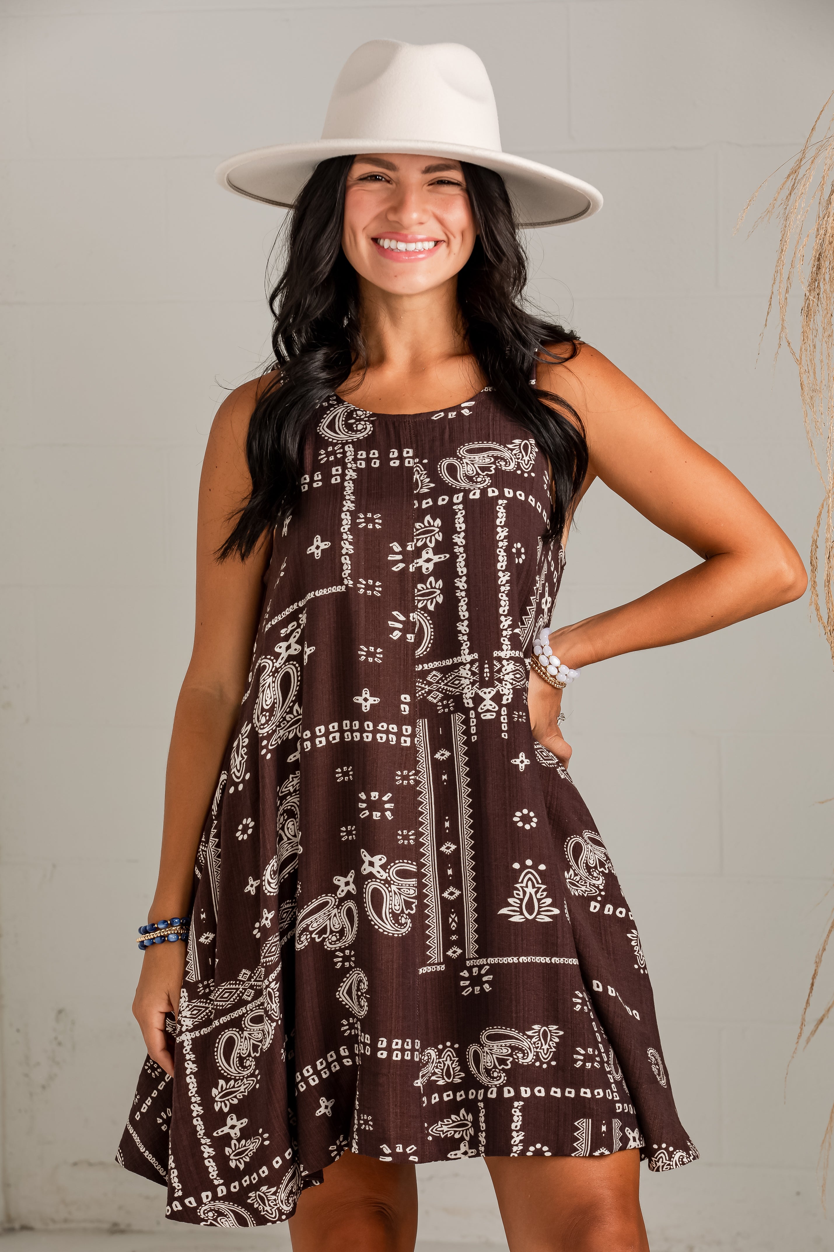 A woman wearing a sleeveless brown dress with a white bandana-style paisley and geometric print, paired with a wide-brimmed white hat, posing against a light gray wall.