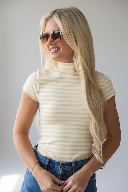 A blonde woman wearing a yellow and white striped mock neck t-shirt and blue jeans, posing against a light gray background.