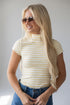 A blonde woman wearing a yellow and white striped mock neck t-shirt and blue jeans, posing against a light gray background.