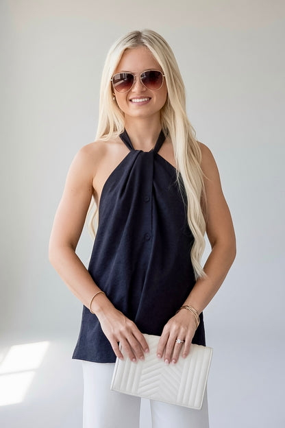 A blonde woman wearing a navy blue halter top, white pants, and sunglasses, holding a white quilted clutch bag.