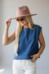 A woman wearing a blue sleeveless top, white pants, a dusty pink wide-brim hat, and sunglasses, posing against a light gray background.