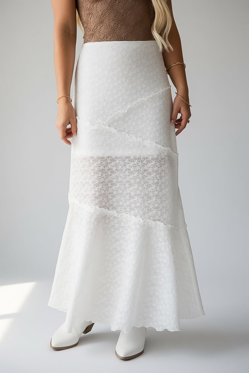A woman wearing a white textured lace maxi skirt with a ruffled hem, paired with white pointed-toe boots against a light gray studio background.