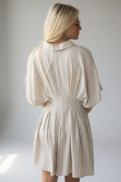A woman with blonde hair is shown from the back wearing a pleated, short dress with wide three-quarter sleeves. The dress is a light cream or off-white color with thin vertical stripes, set against a plain light gray studio background.