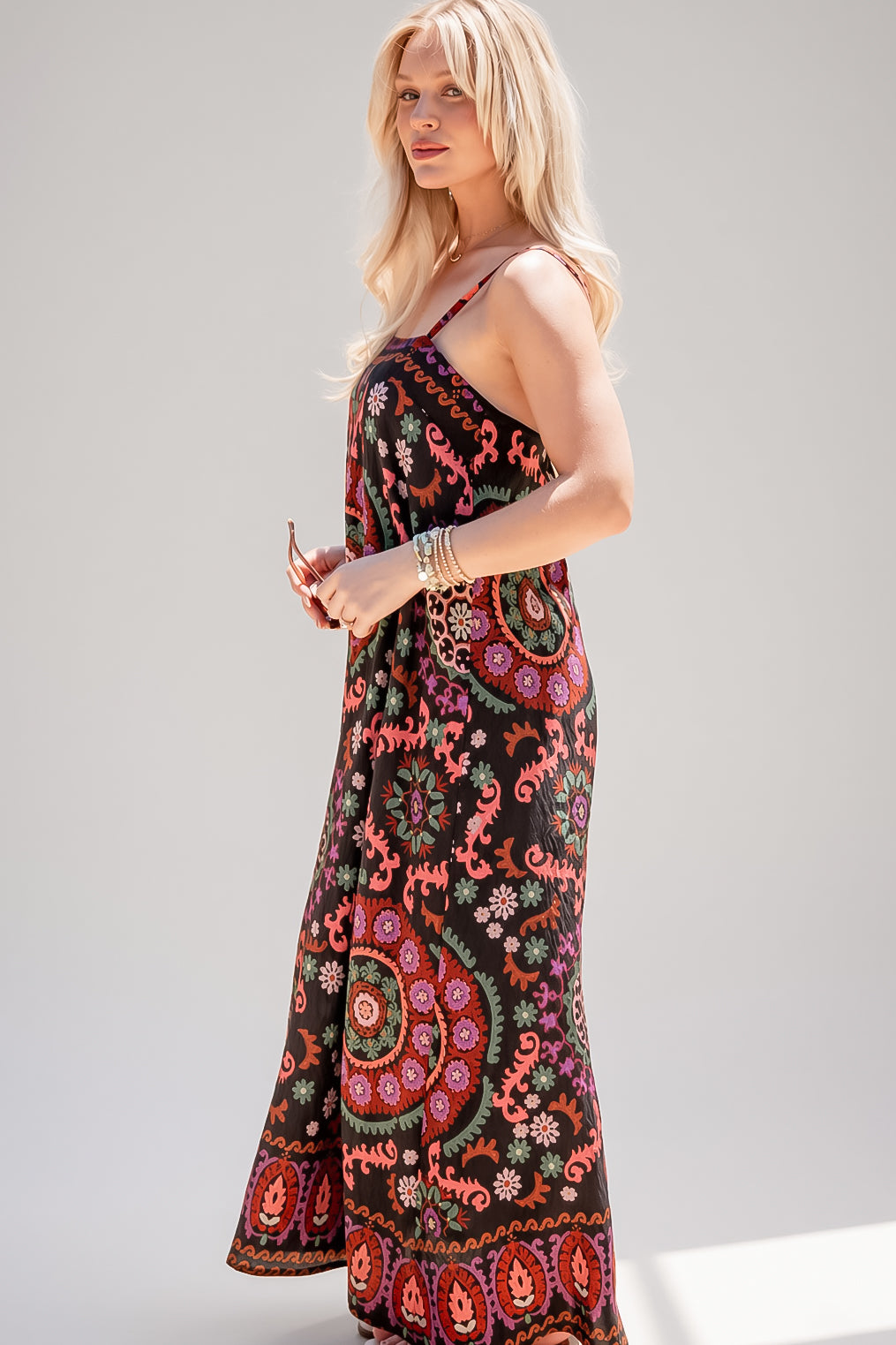 A woman modeling a sleeveless maxi dress with a black base and a vibrant, multicolored bohemian paisley and floral pattern, set against a plain light gray studio background.